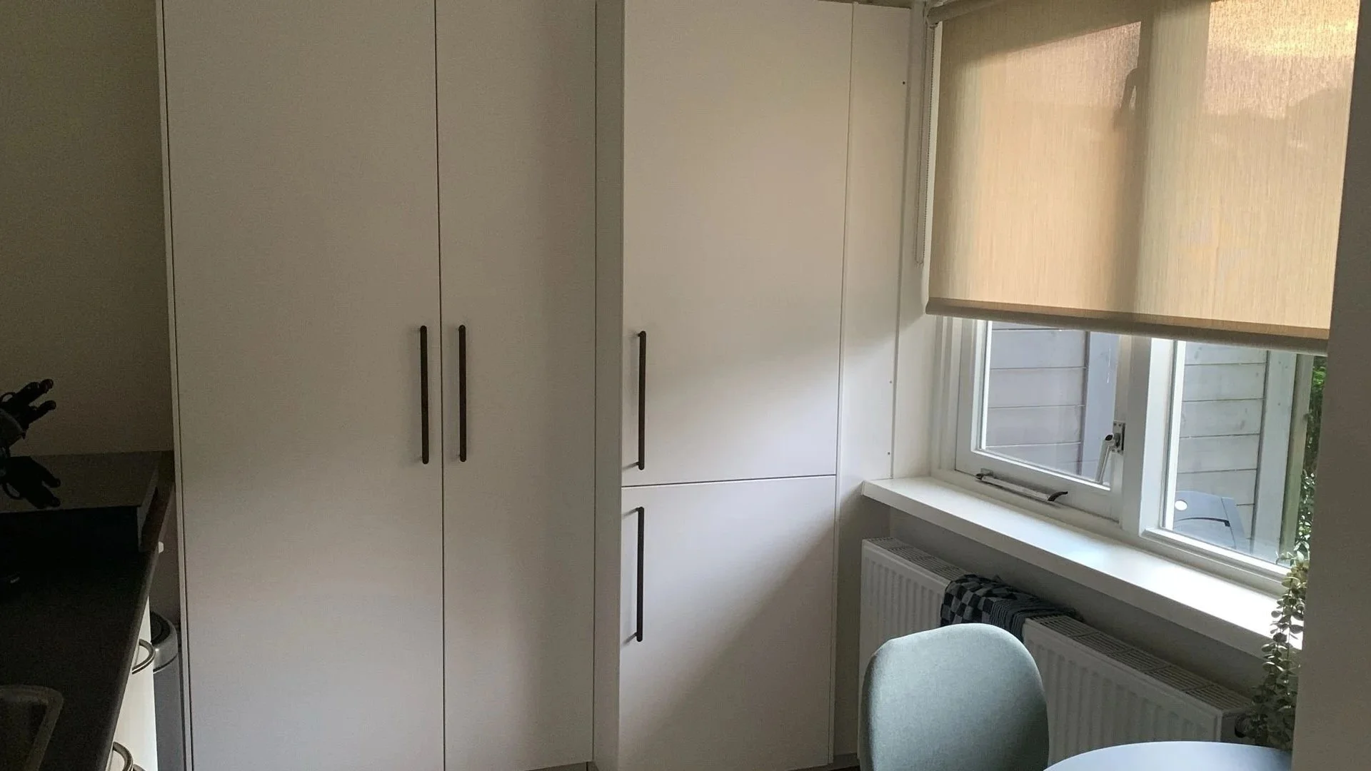 Witkeukenkastje met zwarte handvaten naast een raam met een beige rolgordijn in een keukenruimte.