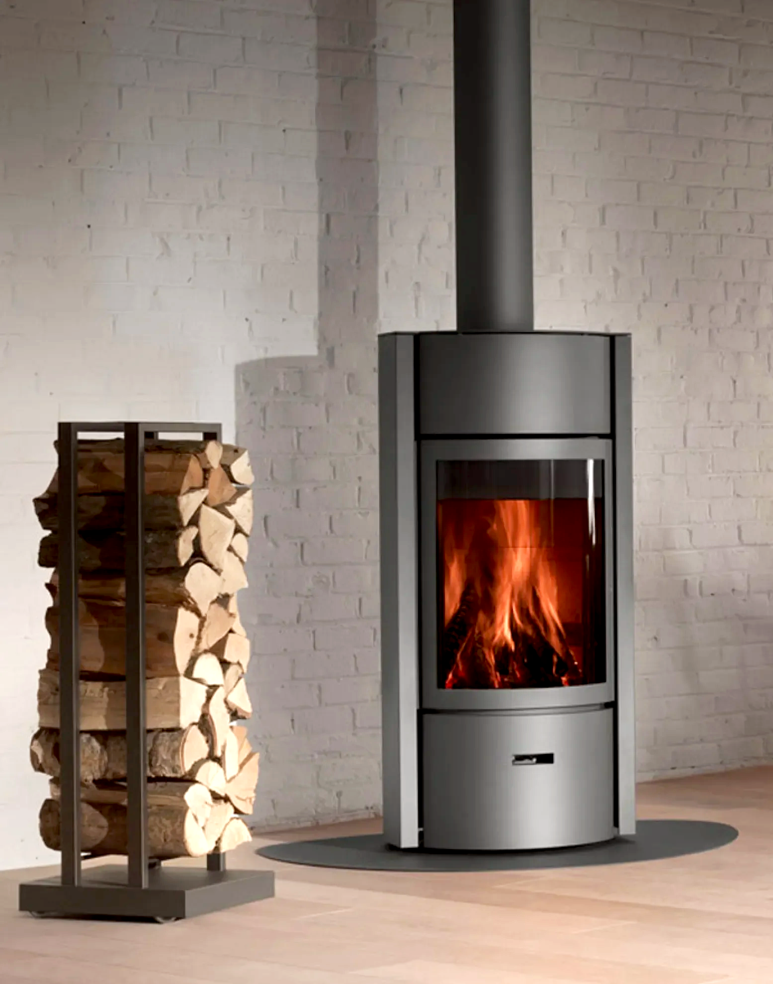 Een moderne houtkachel met een zwarte buis en een houtrek met opgeslagen hout naast een witte bakstenen muur.