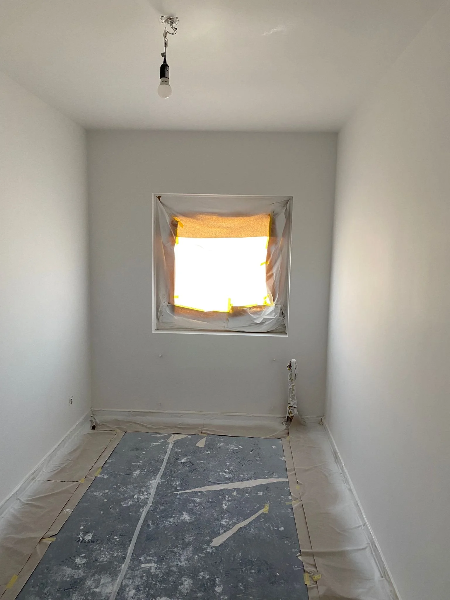 Een slaapkamer onder renovatie, gesloten raam met afgeplakt glas, witte muren, en de vloer afgewerkt met beschermfolie.