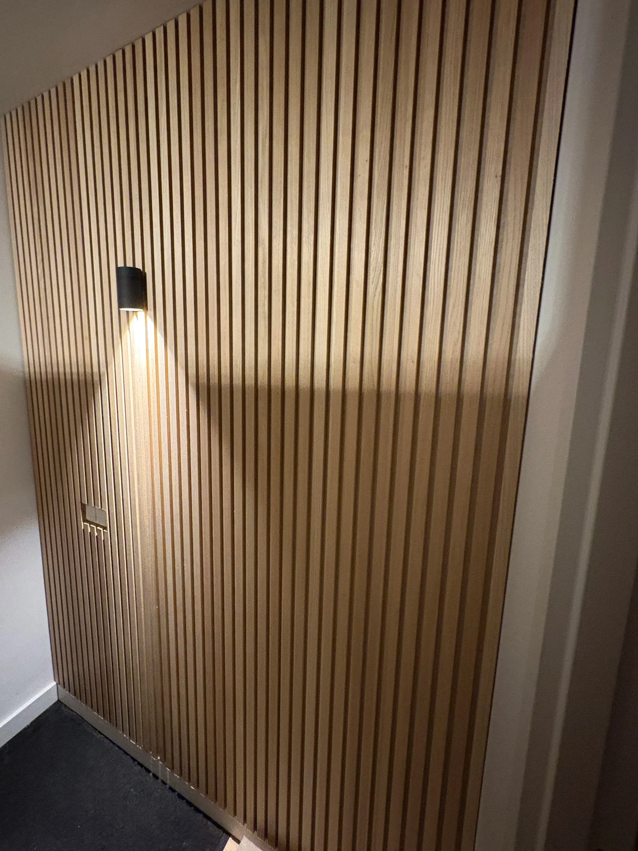 Houtwand met verticale latten en een wandlamp die licht naar beneden werpt. Klusbedrijf.