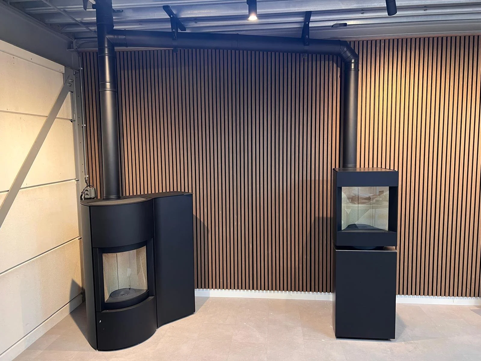 Moderne houtkachel met zwarte afwerking en vaste stenen basis, aangesloten op een zwart rookkanaal, geplaatst voor een houten lamellenmuur in een interieur.