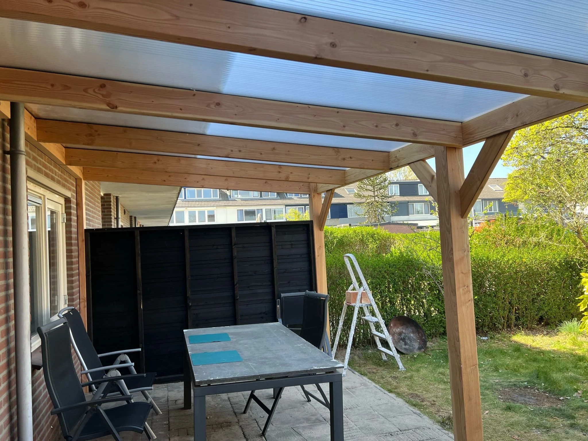 Een beschut terras met een houten afdak, een tafel met stoelen, een ladder en een tuinschijf, omgeven door groen struikgewas en een gebouw op de achtergrond.