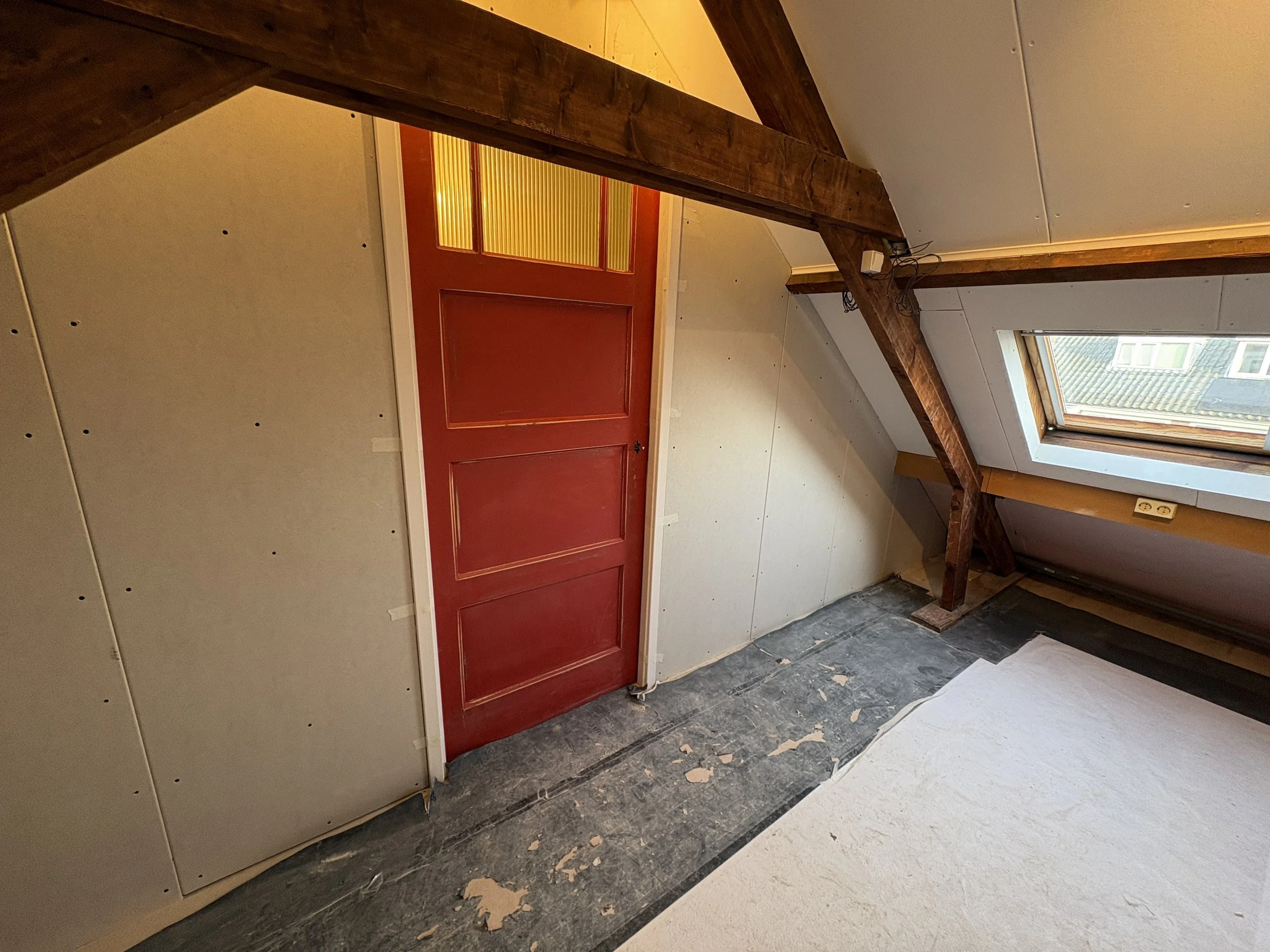 Een zolderruimte met een rode houten deur, een dakraam dat uitzicht biedt op omliggende daken, en onafgewerkte muren met schroefgaten.