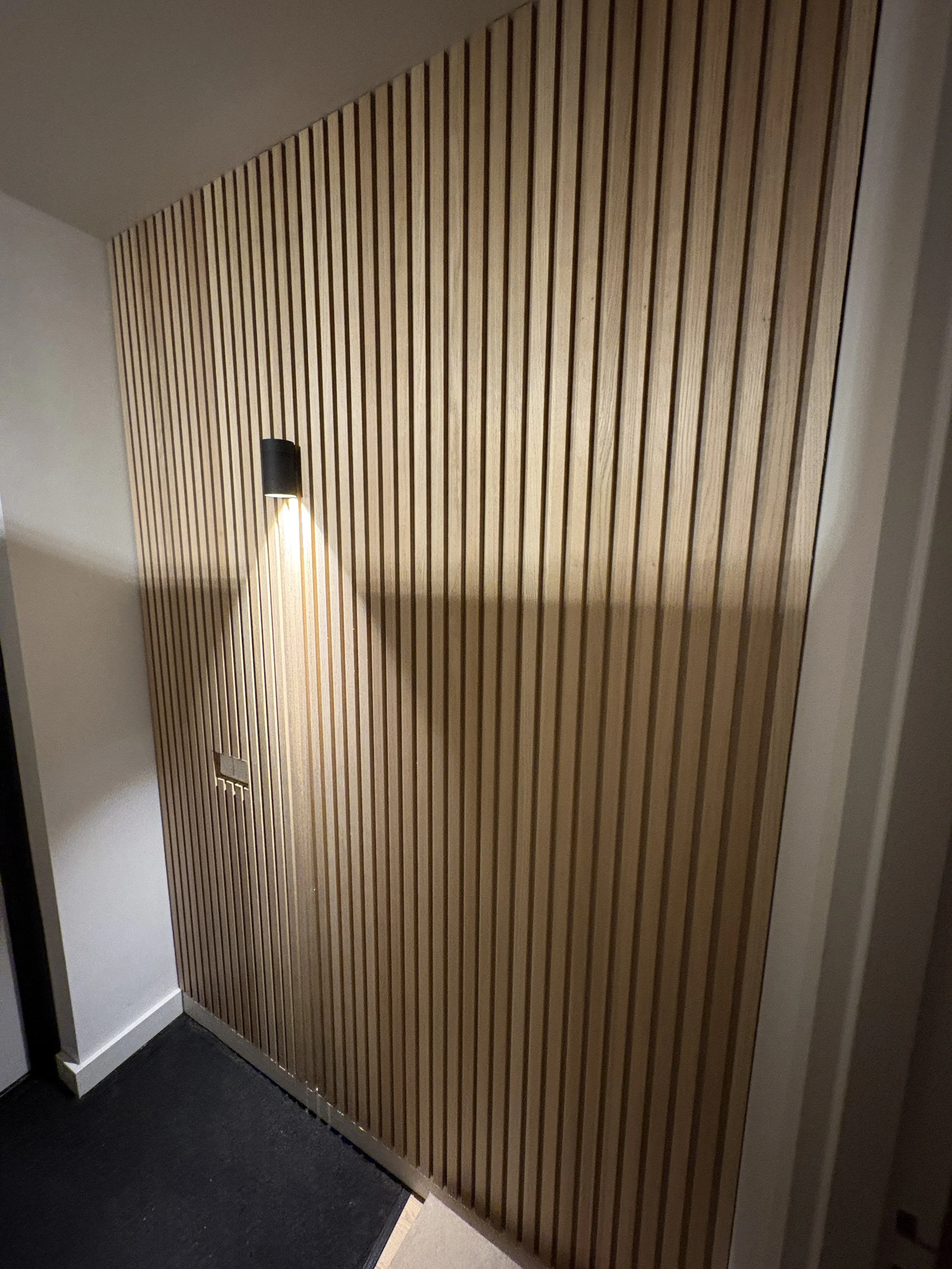 Een muur met verticale houten latten en een zwarte wandlamp die licht uitstraalt.