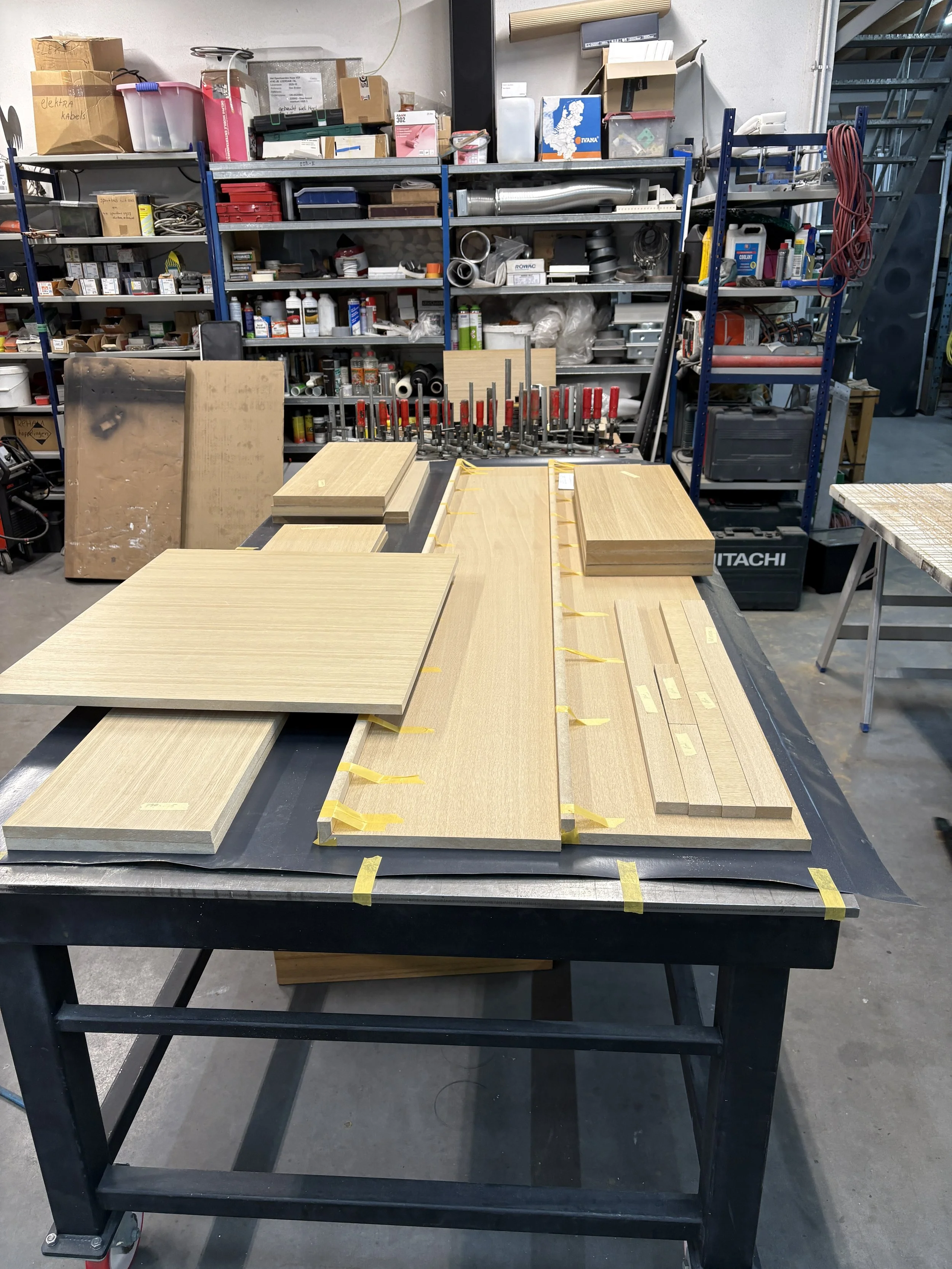 Werkbank met houten meubelpanelen en lijm, in een houtbewerkingswerkplaats met gereedschap en materialen op de achtergrond.