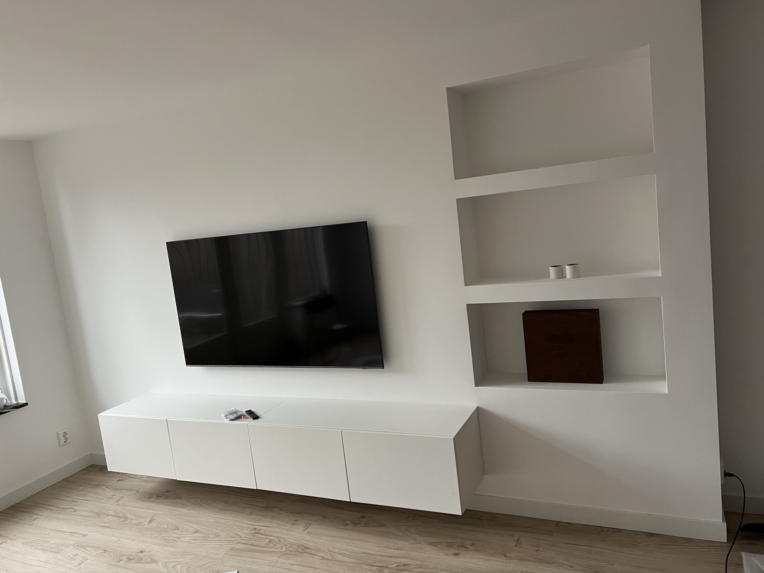 Wit woonkamer met zwarte televisie en ingebouwde witte muurrek. Zwarte tv op witte wandkast met drie deuren, houten vloer, en gekleurde decoraties in de nis.