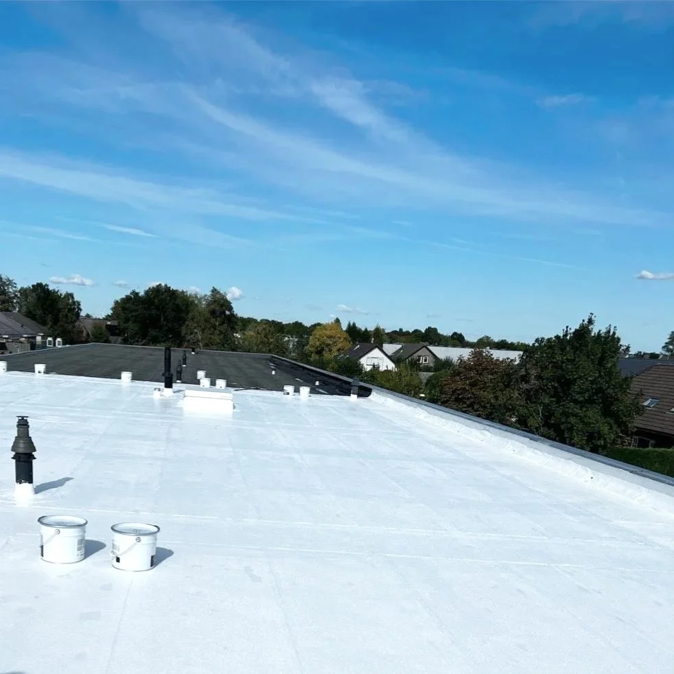 Daken bedekt met witte waterdichte coating, met ventilatiepijpen en dakluchten, onder een blauwe hemel met wolken.