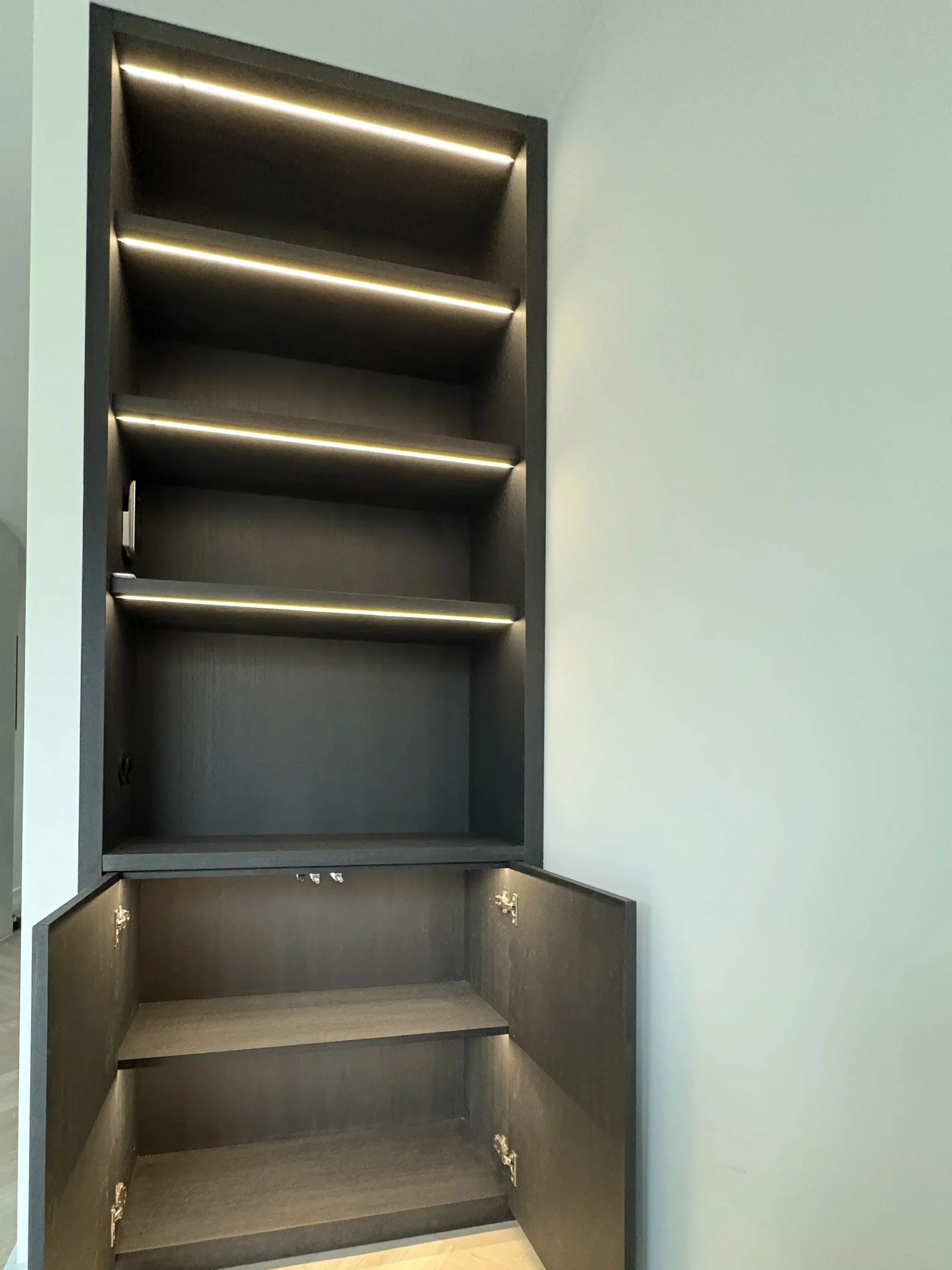 Een verticale zwarte meubelkast met meerdere open planken en een onderliggende compartiment met deuren, tegen een lichte muur.