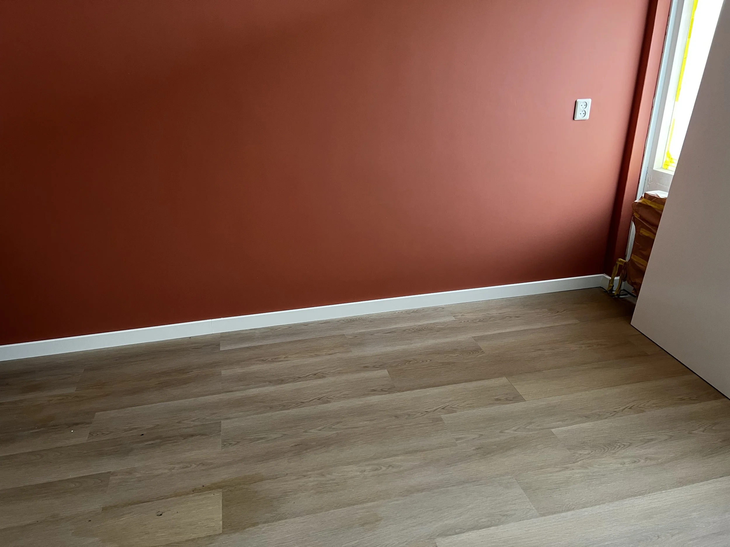 Een kamer met een roodgekleurde muur, een houten vloer en een wit deurlijst. Een raam ligt open en er ligt een geel pakket naast de muur.