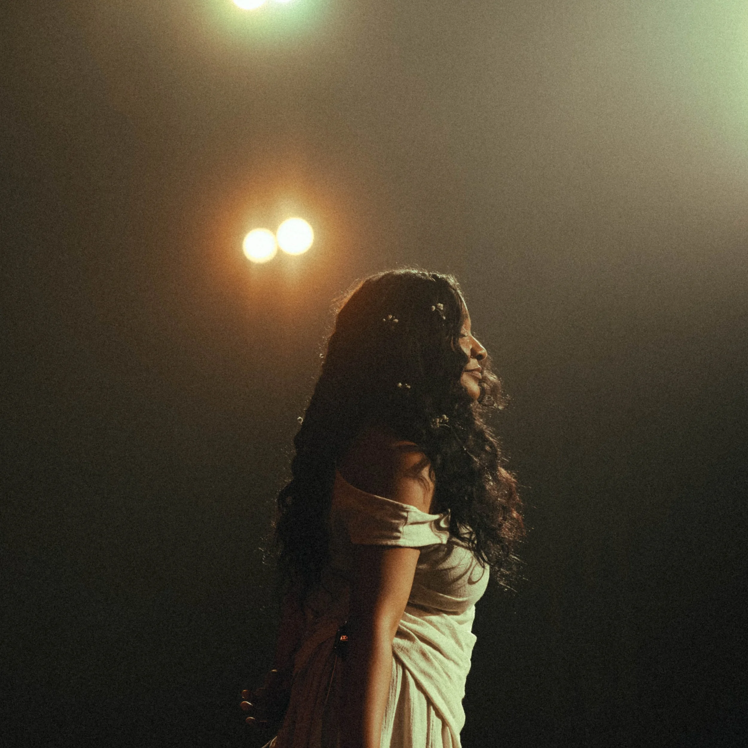 A woman with long, curly hair stands in profile under stage lights.
