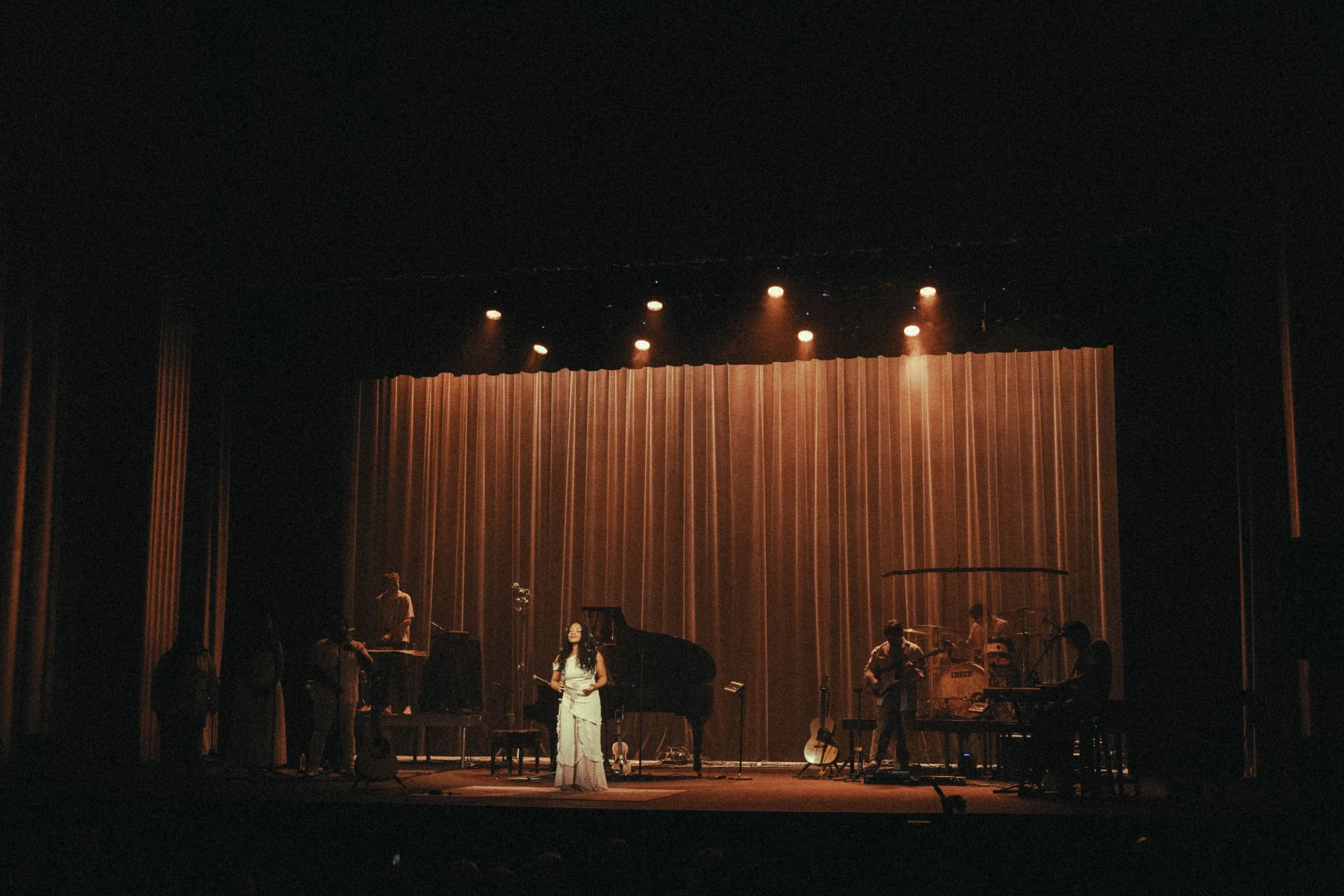 A live music performance on stage with a woman singer in the center, a grand piano, and band members playing guitar, keyboard, and drums under warm stage lights with a brown curtain backdrop.