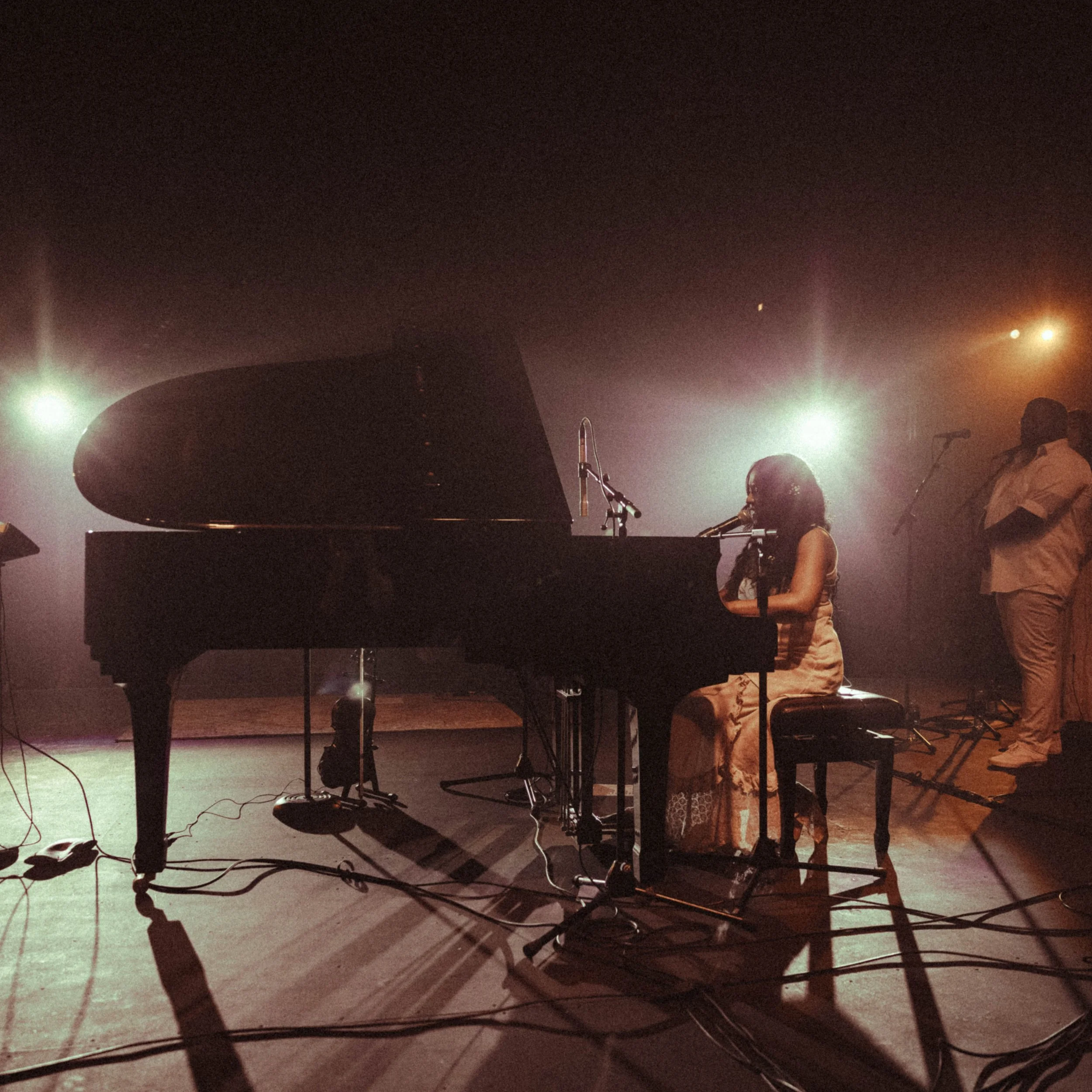 A woman playing a grand piano on a stage with microphone and stage lights shining behind her.