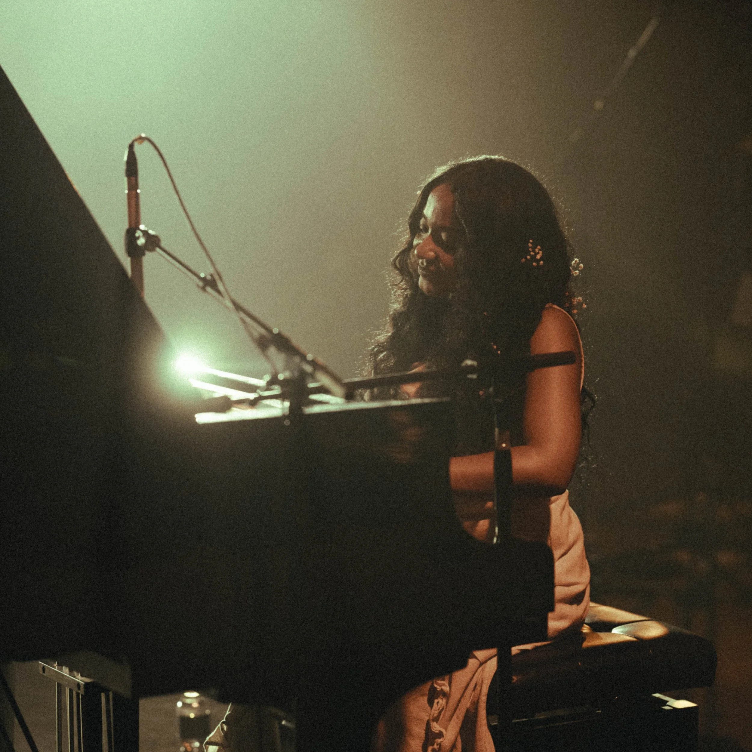 A woman with curly hair and glasses playing a black grand piano on stage, illuminated by soft lighting.
