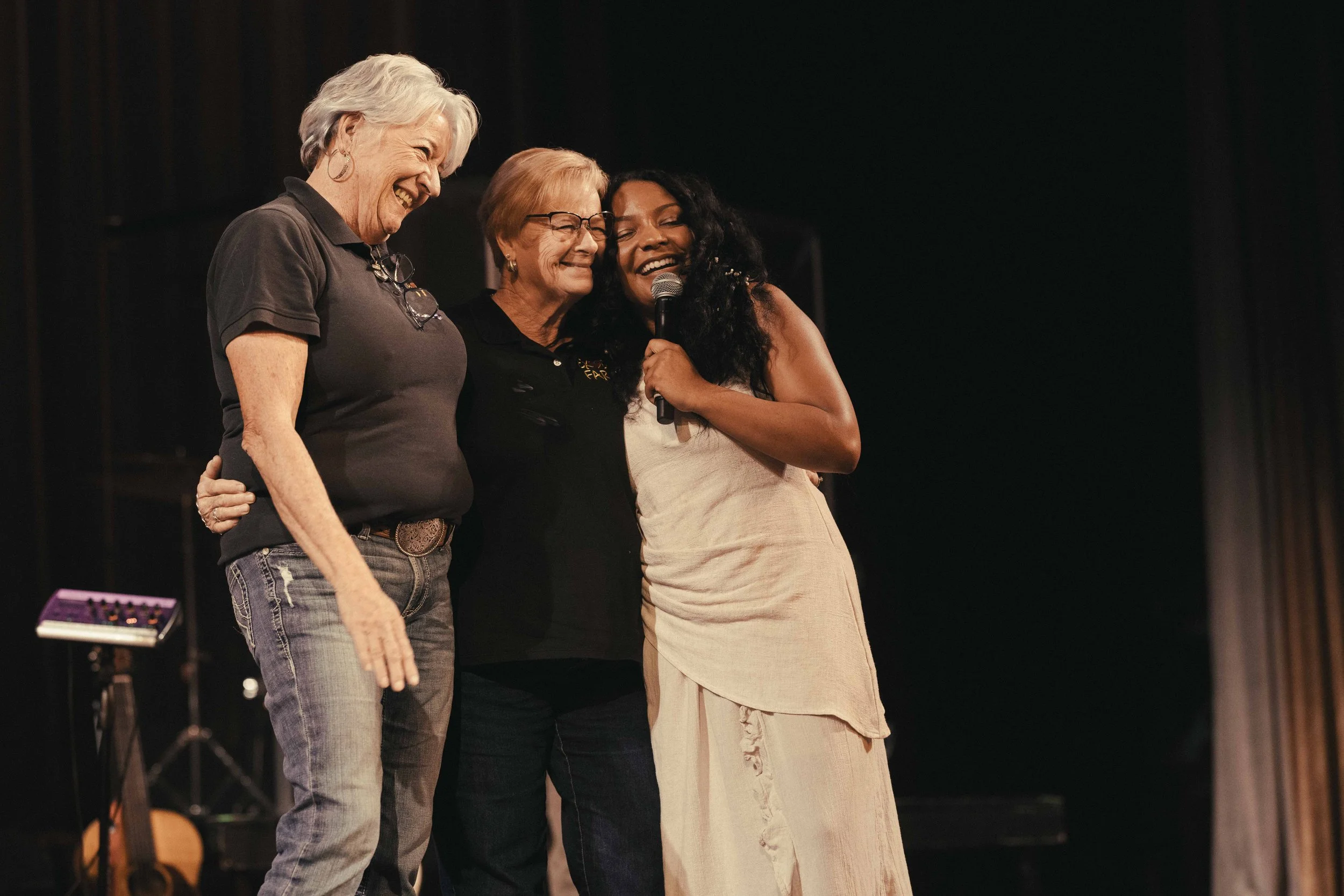 Three women hugging on stage, one holding a microphone, smiling and engaging with audience during a performance or event.