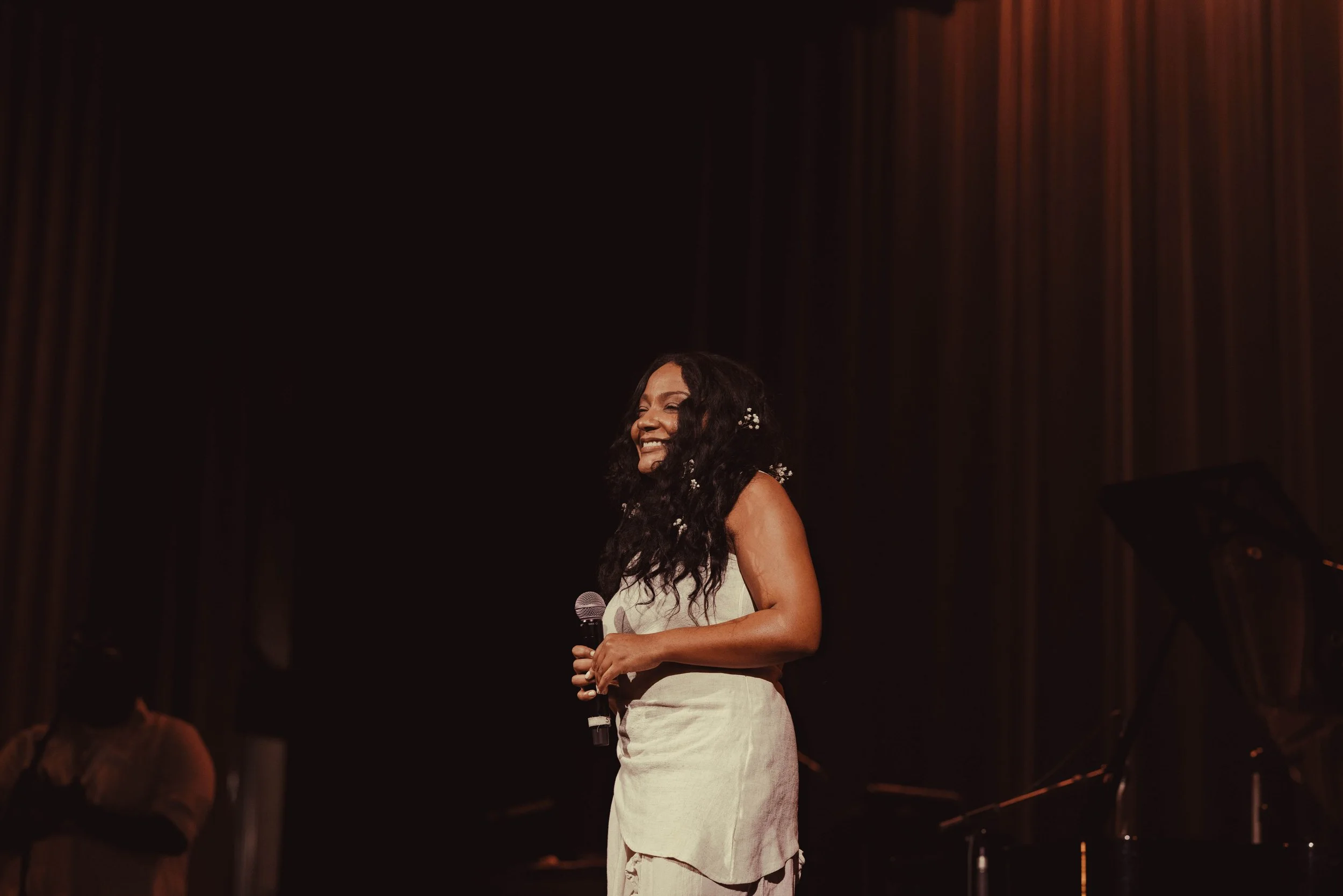 A woman with long, curly black hair smiling and holding a microphone on stage with dark curtains in the background.