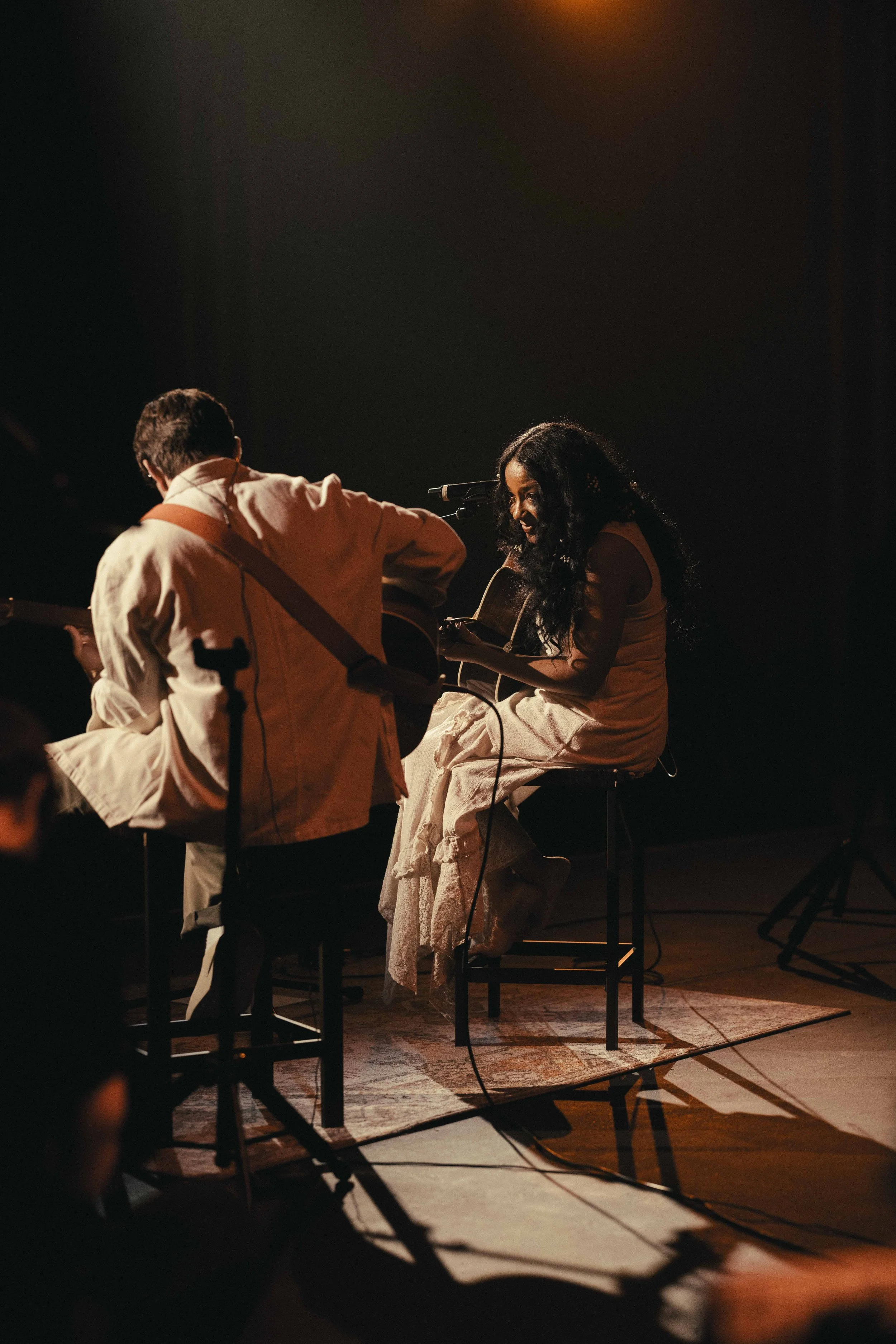 Two musicians performing on stage: a man playing guitar, viewed from the back, and a woman sitting on a chair playing guitar and singing into a microphone. The scene is dimly lit with warm lighting.