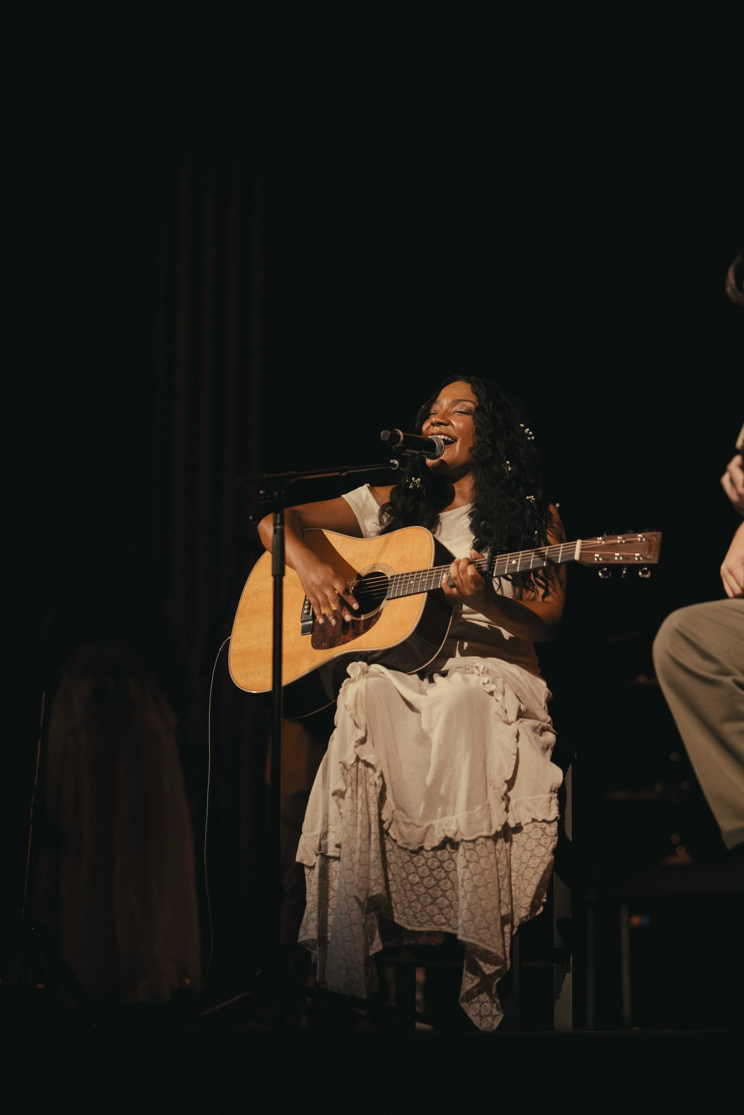 A woman with long black curly hair, wearing a white outfit, is sitting on a stool playing an acoustic guitar and singing into a microphone on stage.