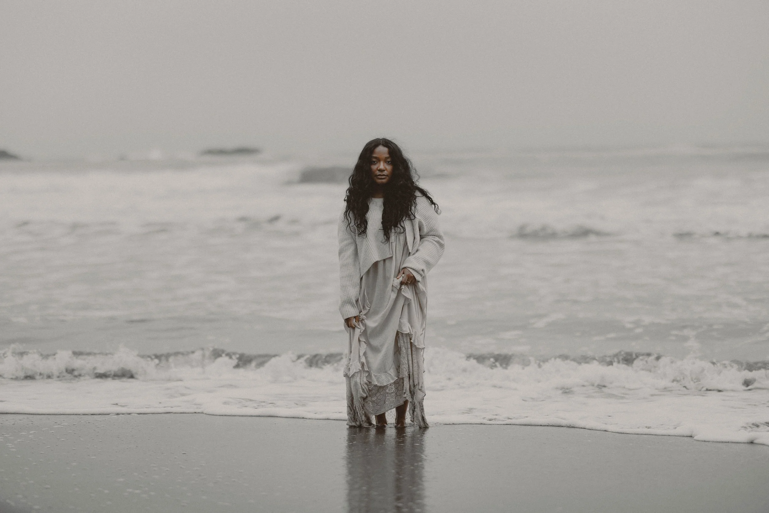 Woman standing in shallow ocean water at the beach on a cloudy day, wearing a flowing dress and sweater.