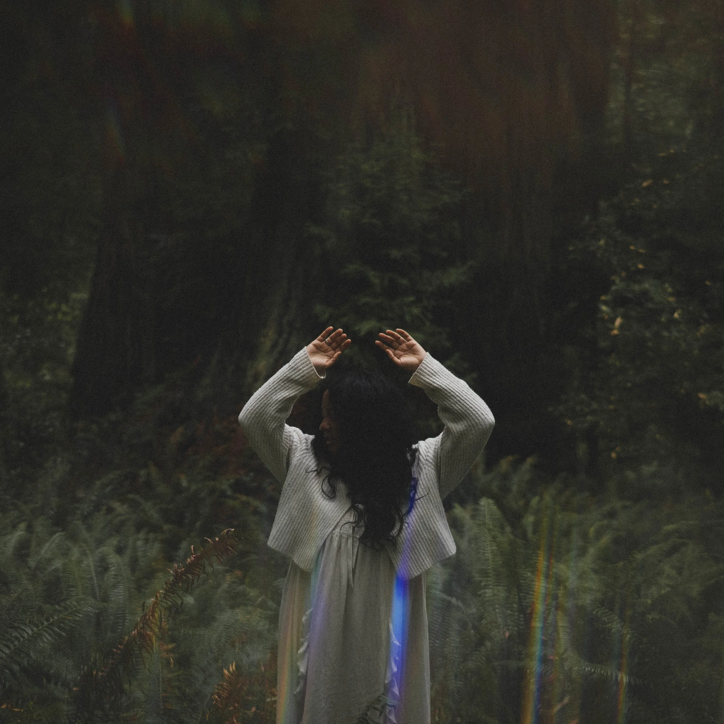 A woman with dark hair and dark skin standing in a forest with tall trees and ferns, raising their arms above their head, wearing a white garment and a knitted cardigan.