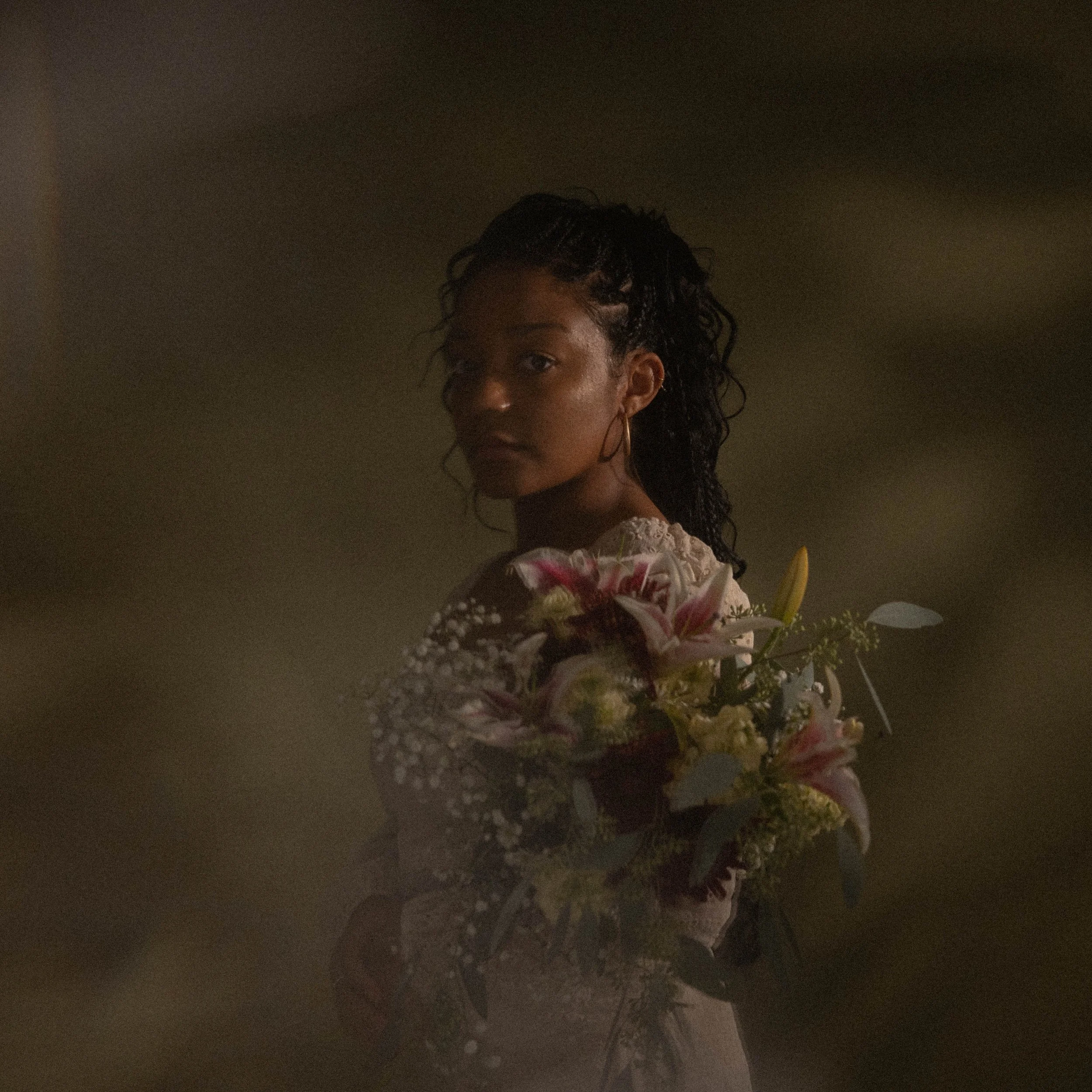 A woman with dark curly hair, wearing a lace dress, holding a bouquet of pink lilies and white flowers, standing in dim lighting.