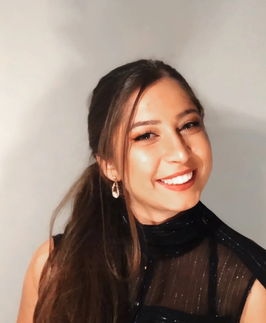 A young woman smiling with long, dark hair, wearing a black sleeveless dress with sheer fabric and sparkles, and dangling earrings, standing against a plain wall.