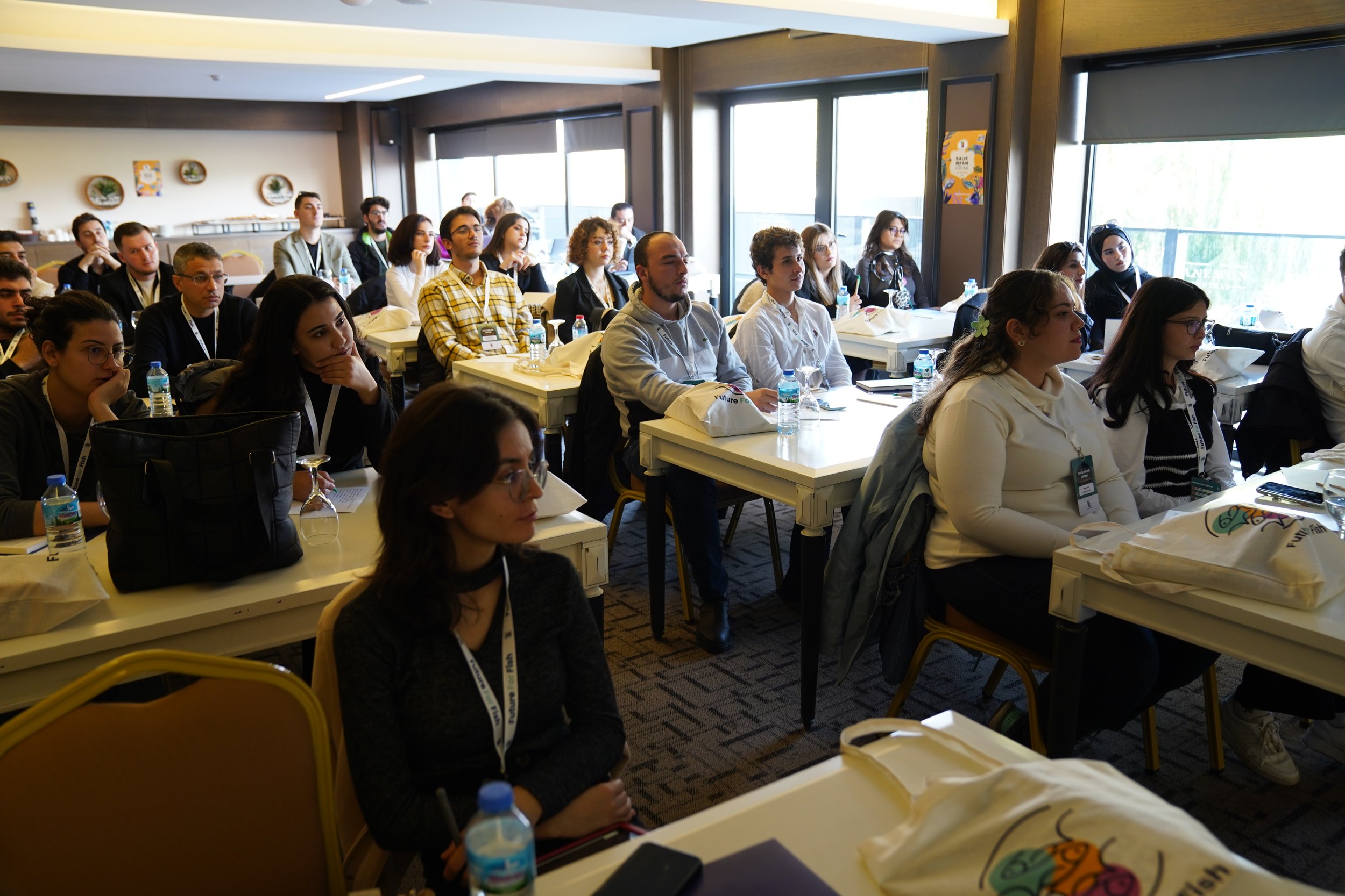 People attending a conference or seminar seated at tables in a bright room with large windows, listening attentively.