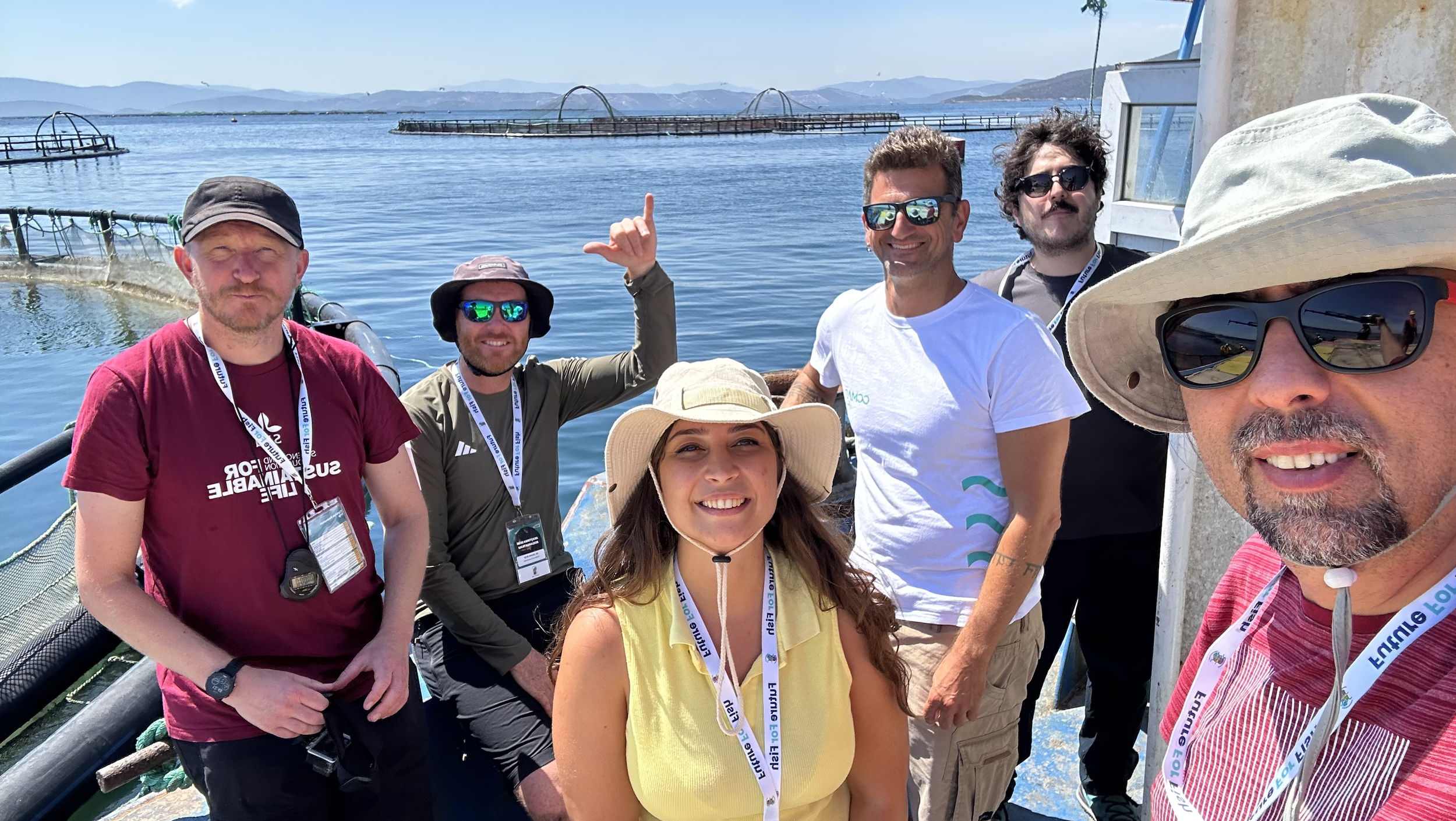 Group of six people on a boat near a lake with mountains in the background, some wearing sunglasses, hats, and casual outdoor clothing.