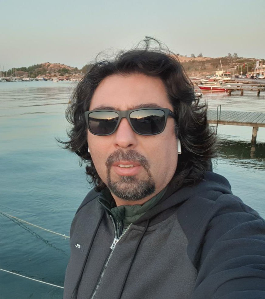 A man with dark curly hair and a beard, wearing sunglasses, AirPods, and a black jacket, taking a selfie by a marina with boats and a hilly landscape in the background.