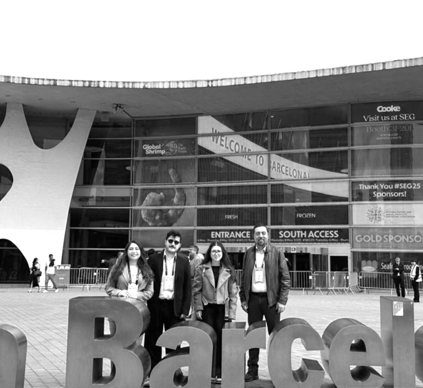 Group of four people standing in front of a large Barcelona sign outside a modern building, with other people walking in the background.