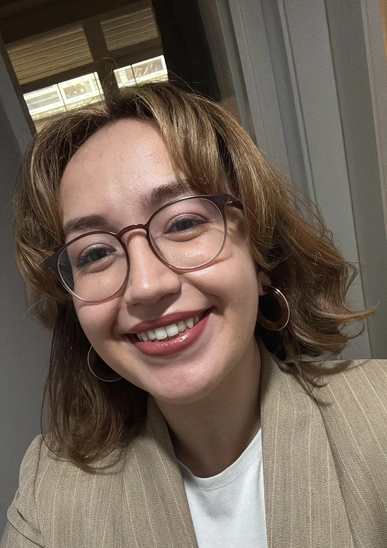 A young woman with light brown, wavy hair wearing glasses, hoop earrings, a beige blazer, and a white shirt, smiling in front of a window.