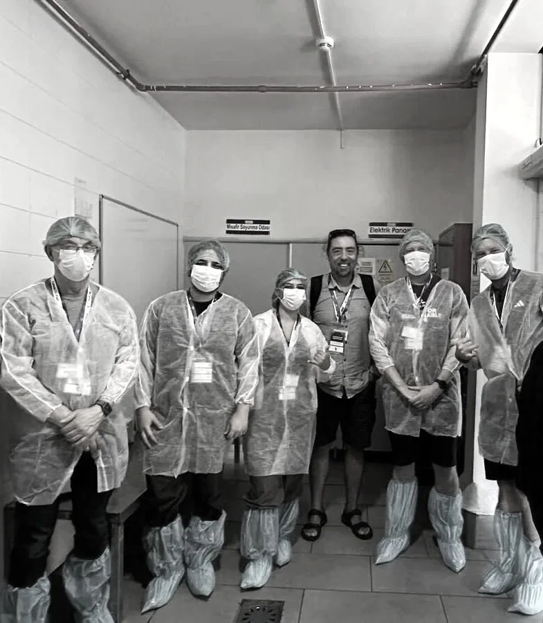 Group of six healthcare workers standing in a corridor, wearing masks, protective gowns, shoe covers, and badges, posing for a photo.