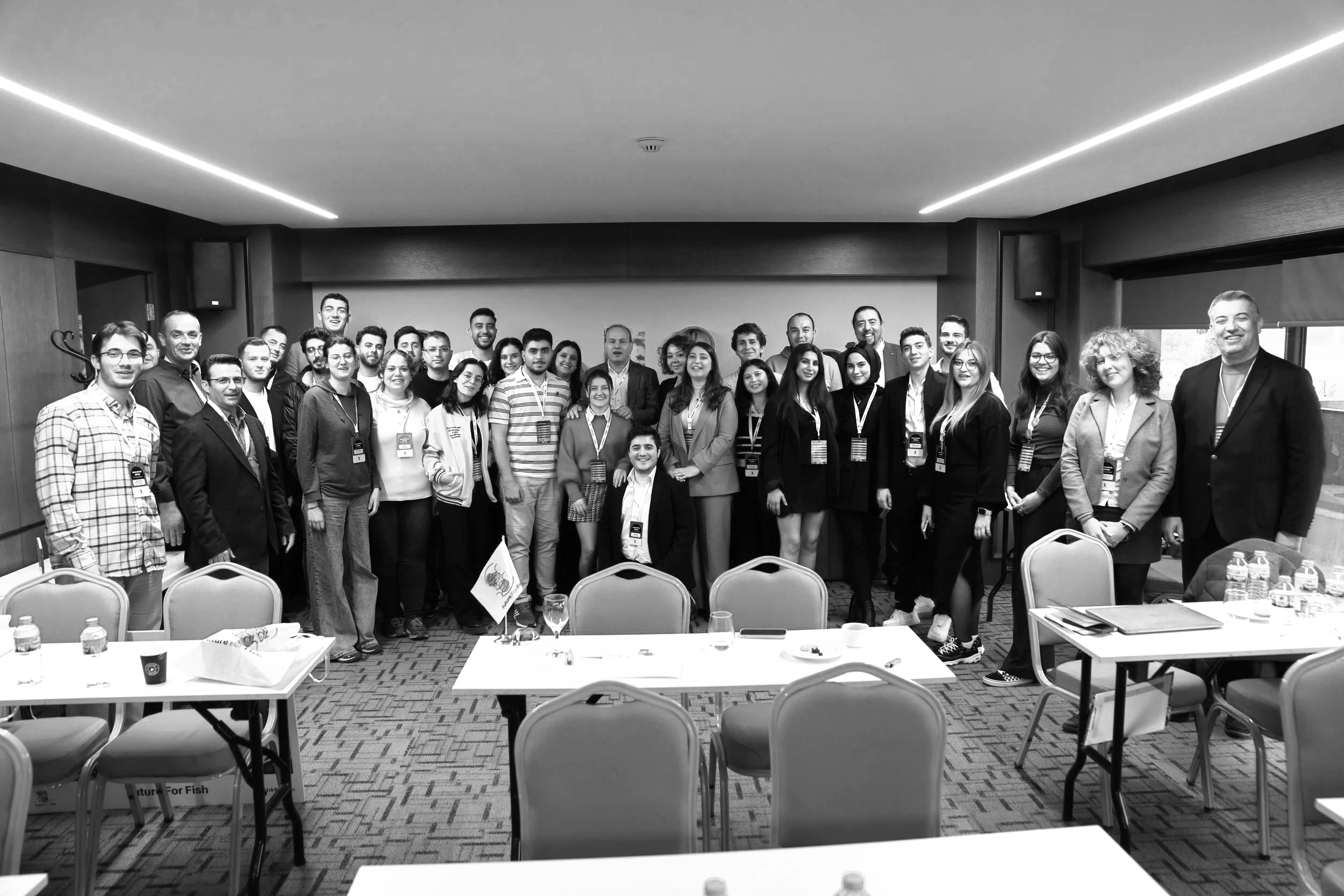 A group of diverse people posing for a photo in a conference room, some wearing conference badges, with tables and chairs in the foreground.