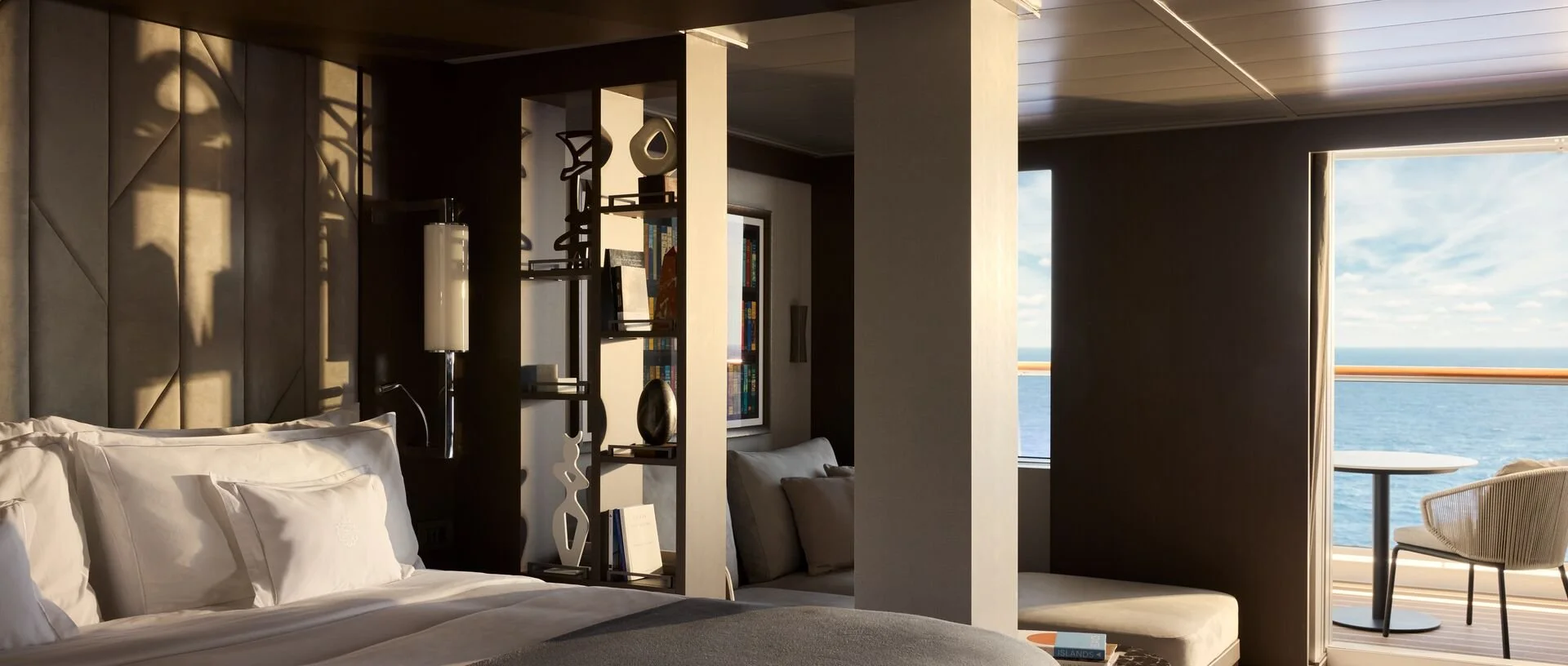 Interior of a hotel room with a bed, bookshelves, a window with ocean view, and outdoor balcony with a table and chair.