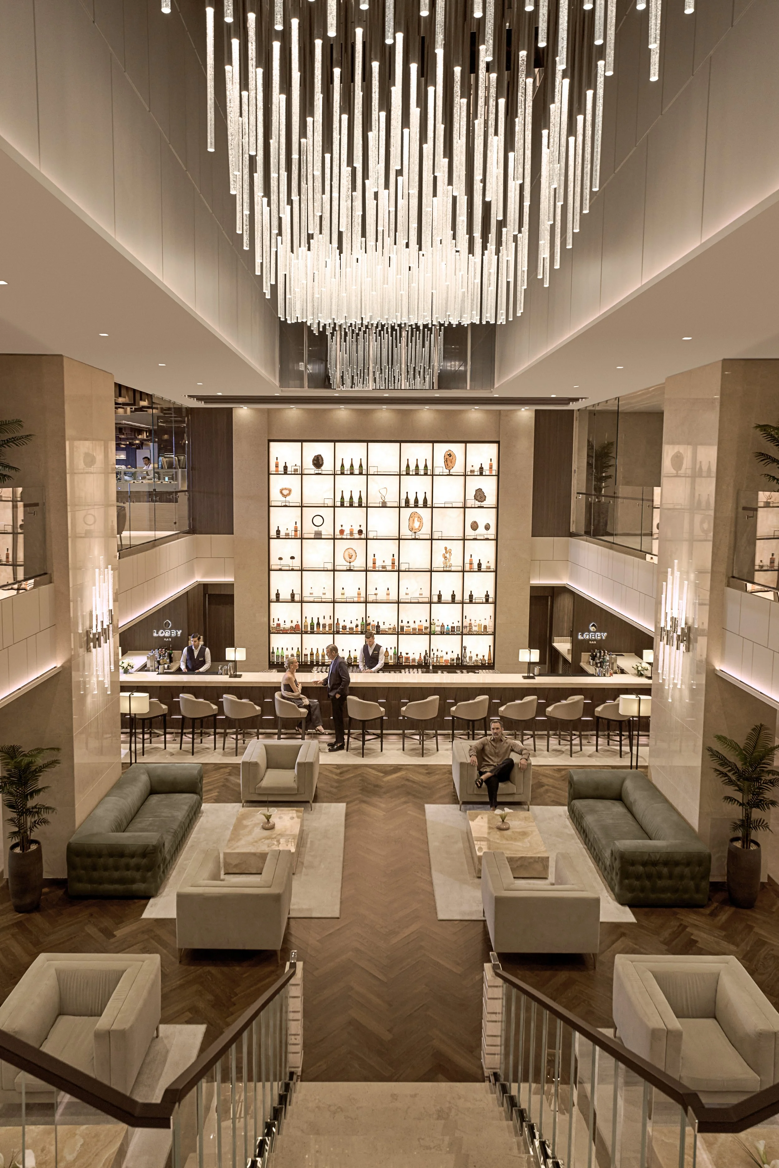 A luxury hotel lobby with a large central bar, a chandelier hanging from the ceiling, and seating areas with sofas and armchairs. The background features a well-lit display of bottles and decorative objects on shelves.