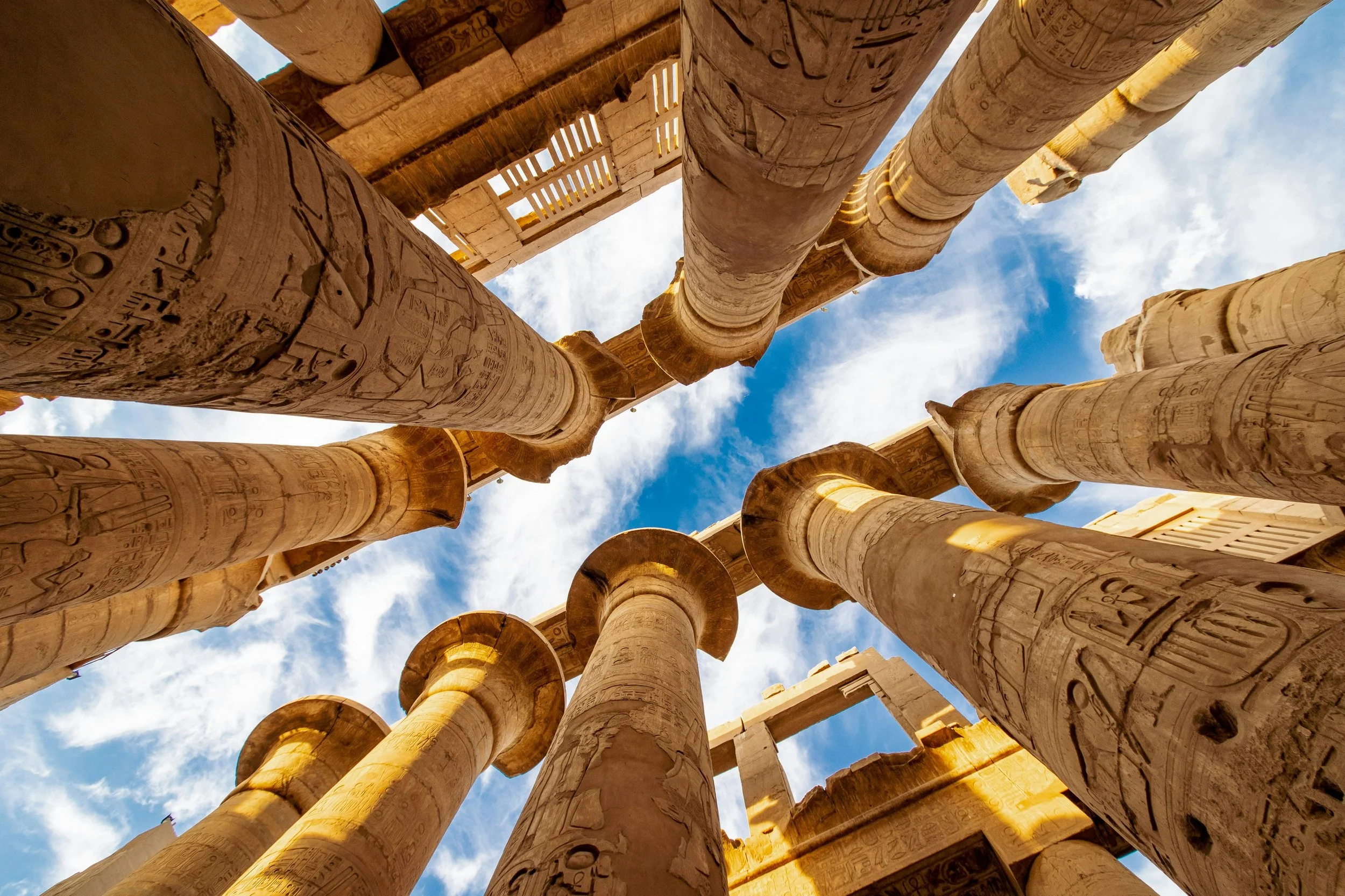 Looking up at the towering carved columns of the Great Hypostyle Hall at Karnak Temple, Luxor, against a blue sky.