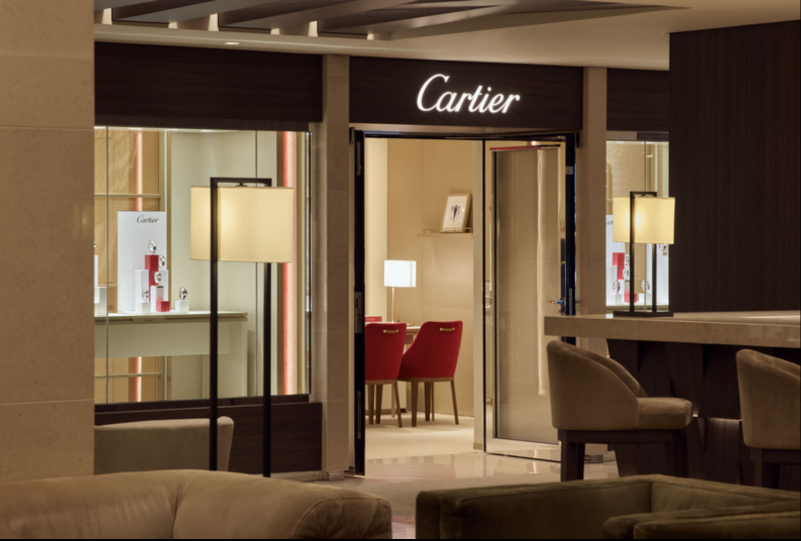 The Cartier store entrance with illuminated sign, glass doors, and interior featuring red chairs, lamps, and display cases with jewelry.