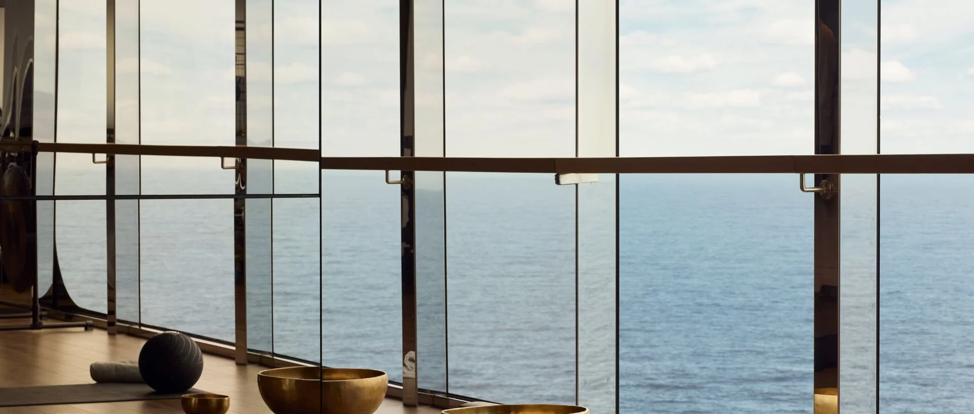 A spacious fitness room with large windows overlooking the ocean, featuring yoga mats, a black exercise ball, and brass bowls on the wooden floor.