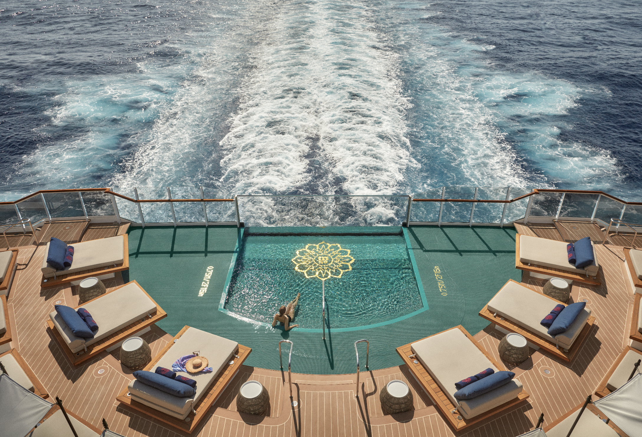 Top view of a cruise ship's deck showing multiple lounge chairs with towels and hats, a small swimming pool, and the ocean water behind the ship creating a wake.
