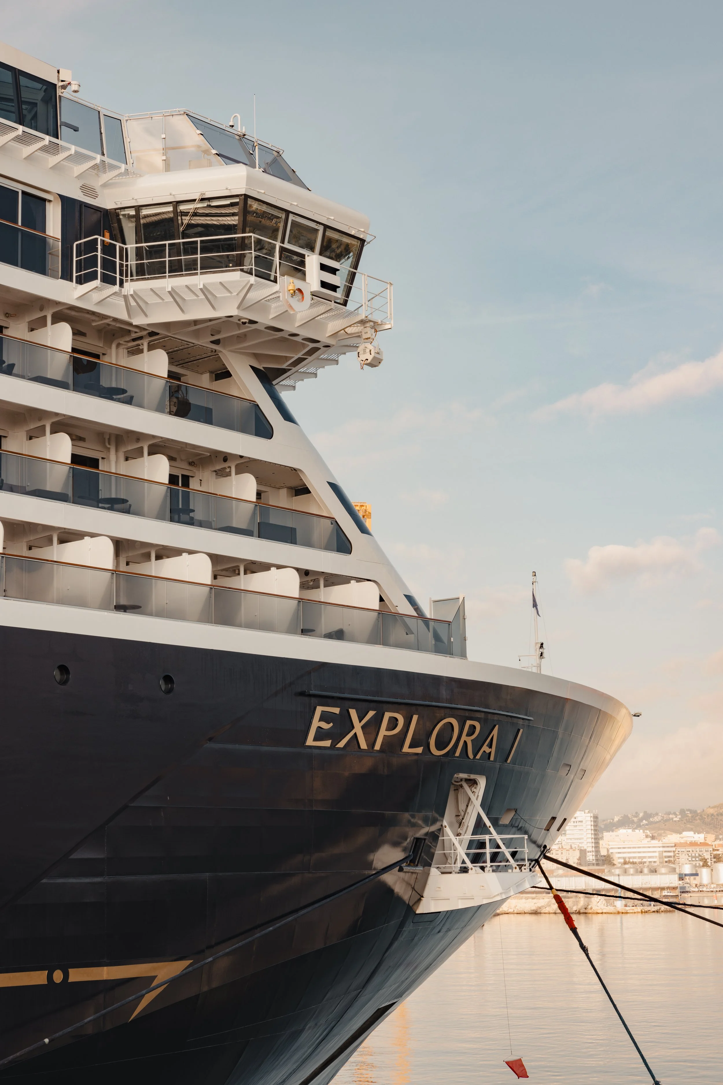 Close-up of the bow of a large cruise ship named EXPLORA, docked at a harbor with city buildings in the background, during daytime with partly cloudy skies.