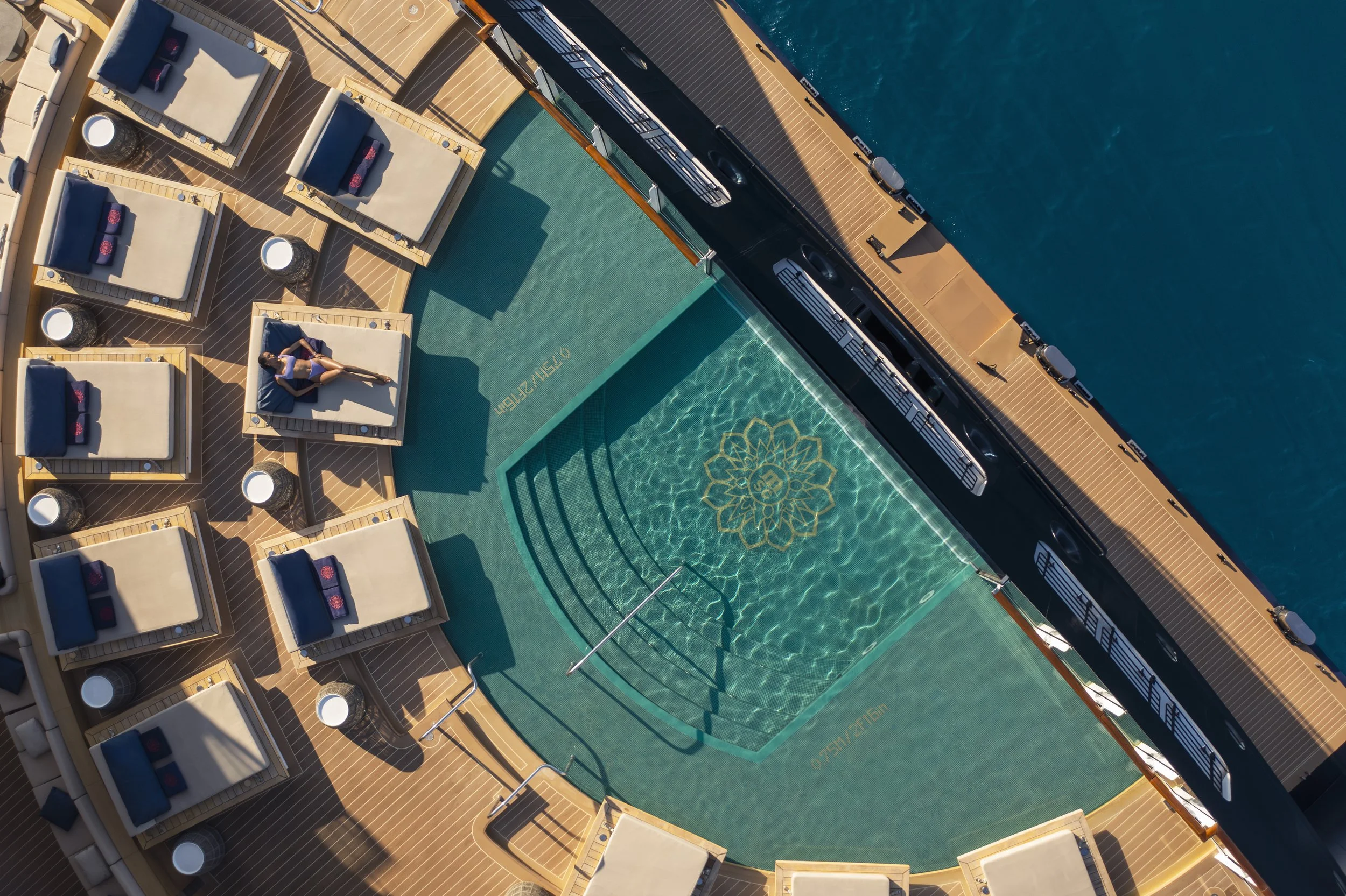 Top view of a cruise ship deck showing a small swimming pool with a decorative flower mosaic and surrounding sun loungers with navy blue cushions and red towels.