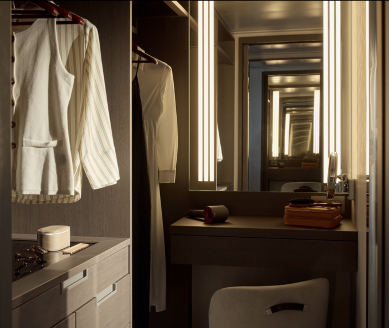 A bathroom vanity area with illuminated mirrors, hanging clothes, and various personal items on the counter.