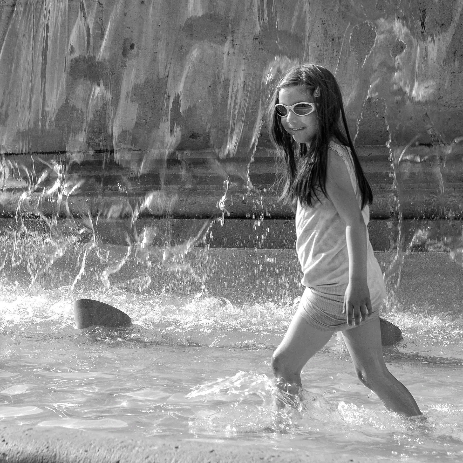 Child running through fountain, sunglasses on, midday
