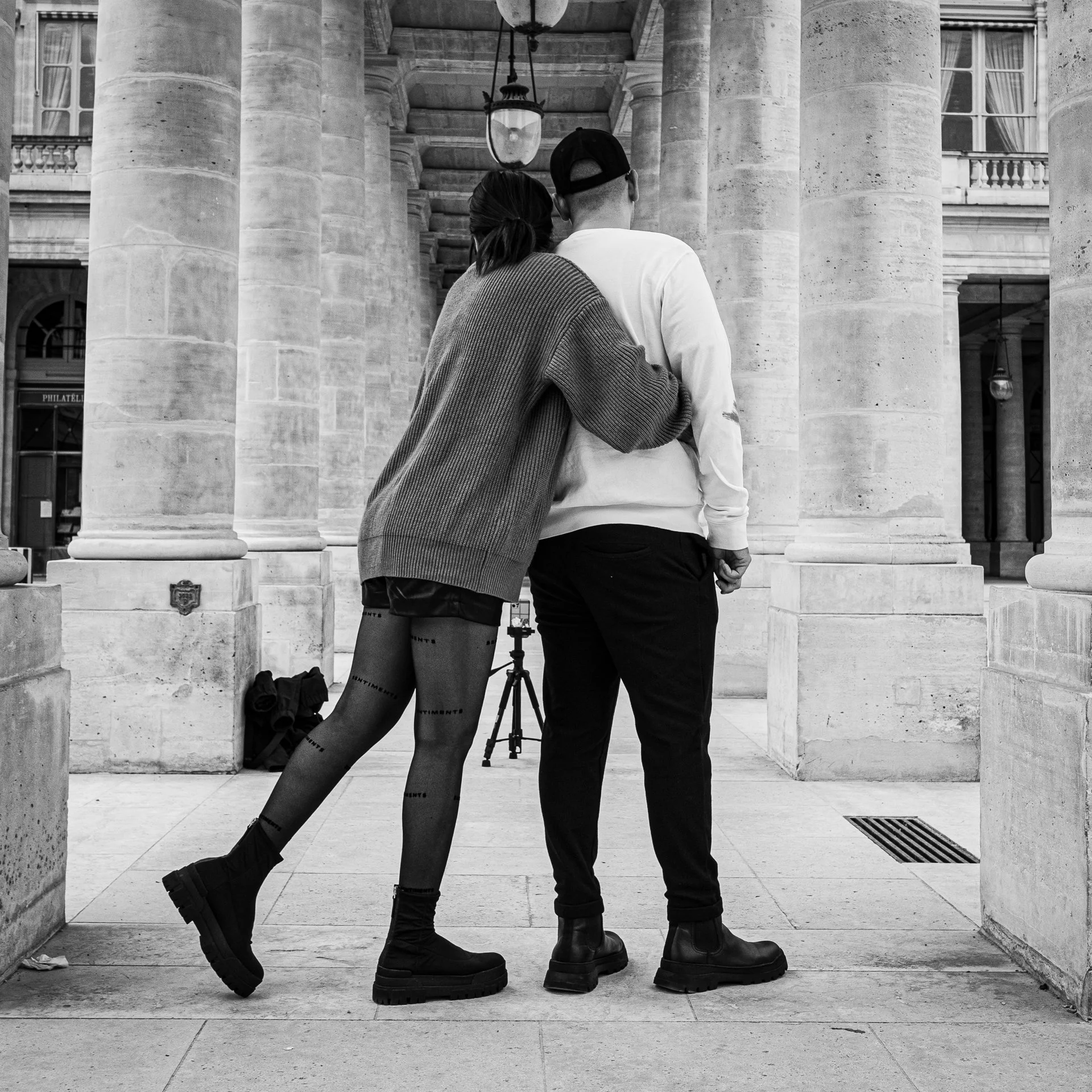 A photograph shows a couple standing close together in a grand colonnade, leaning toward each other as they prepare to take a selfie. Tall stone columns and hanging lamps frame the scene, and a small tripod rests on the ground behind them.