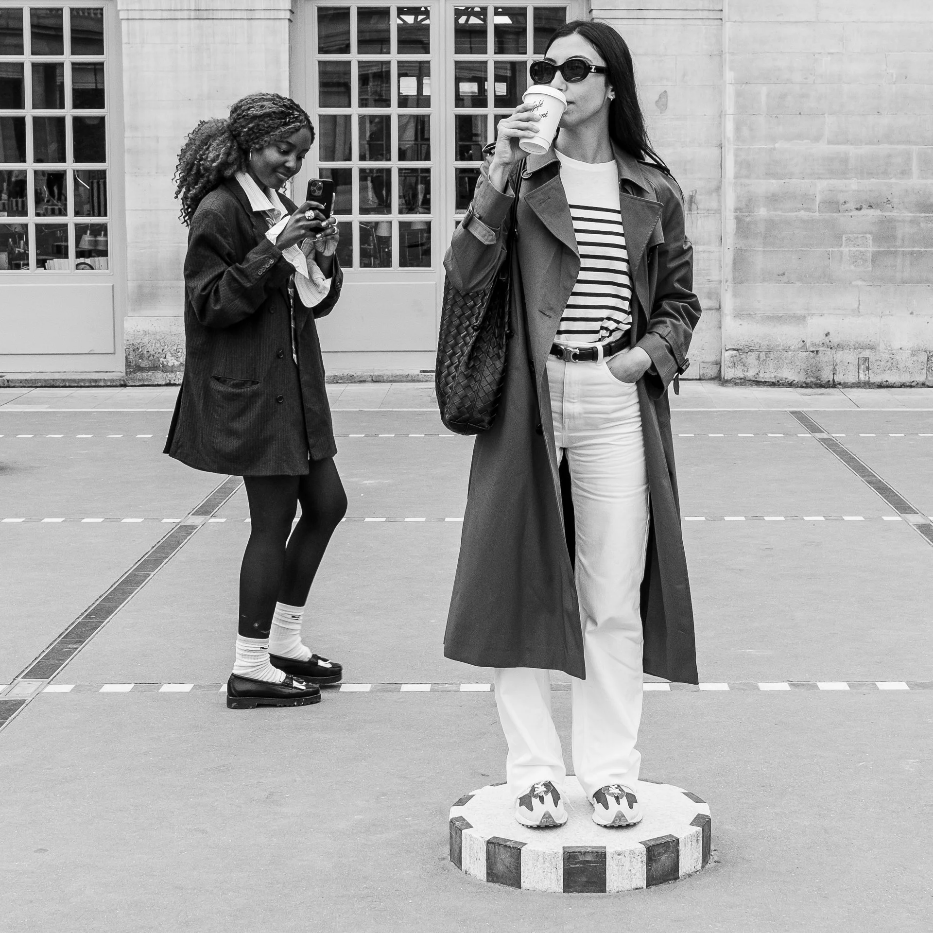 Two figures in black and white: a trench‑coated sitter on a striped pedestal and a companion absorbed in a phone, the plaza’s geometry framing a quiet urban duet.