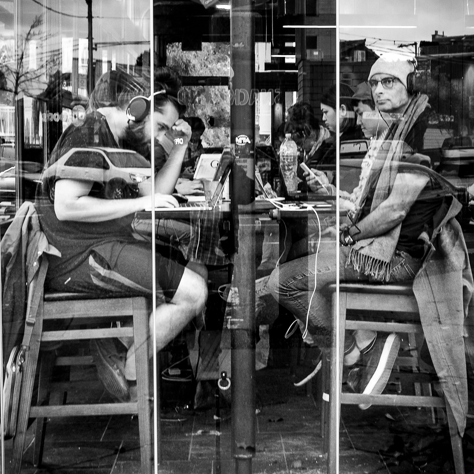 A black‑and‑white photograph taken through a café window shows several people seated at a long table, each absorbed in laptops, phones, or headphones.