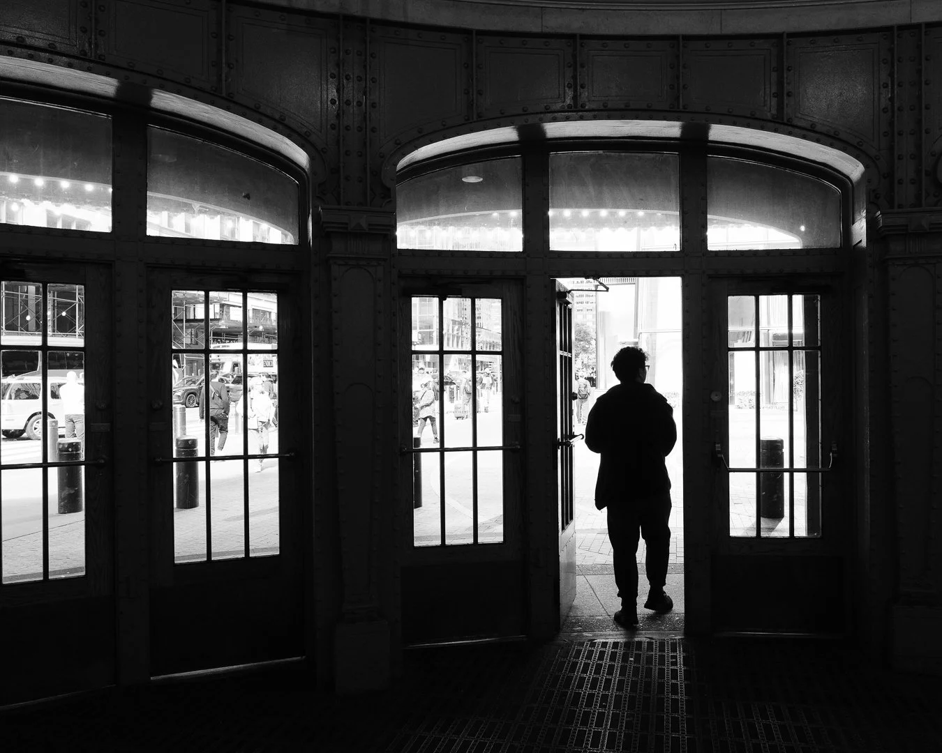 Black-and-white silhouette of a person walking through glass double doors framed by ornate metalwork, bright street scene beyond with pedestrians and vehicles.