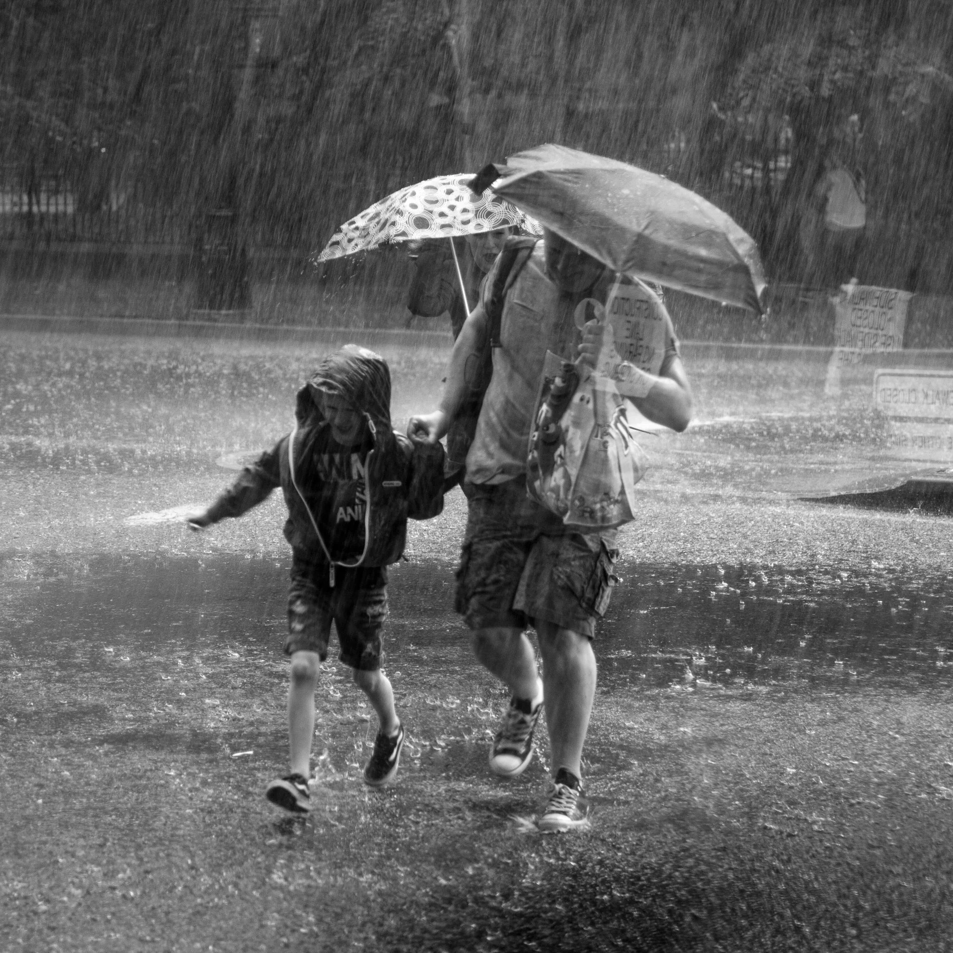 A black‑and‑white street photograph of two figures hurrying through heavy rain. An adult holds a patterned umbrella while a child in a hooded jacket runs close beside them. Raindrops streak diagonally across the frame.
