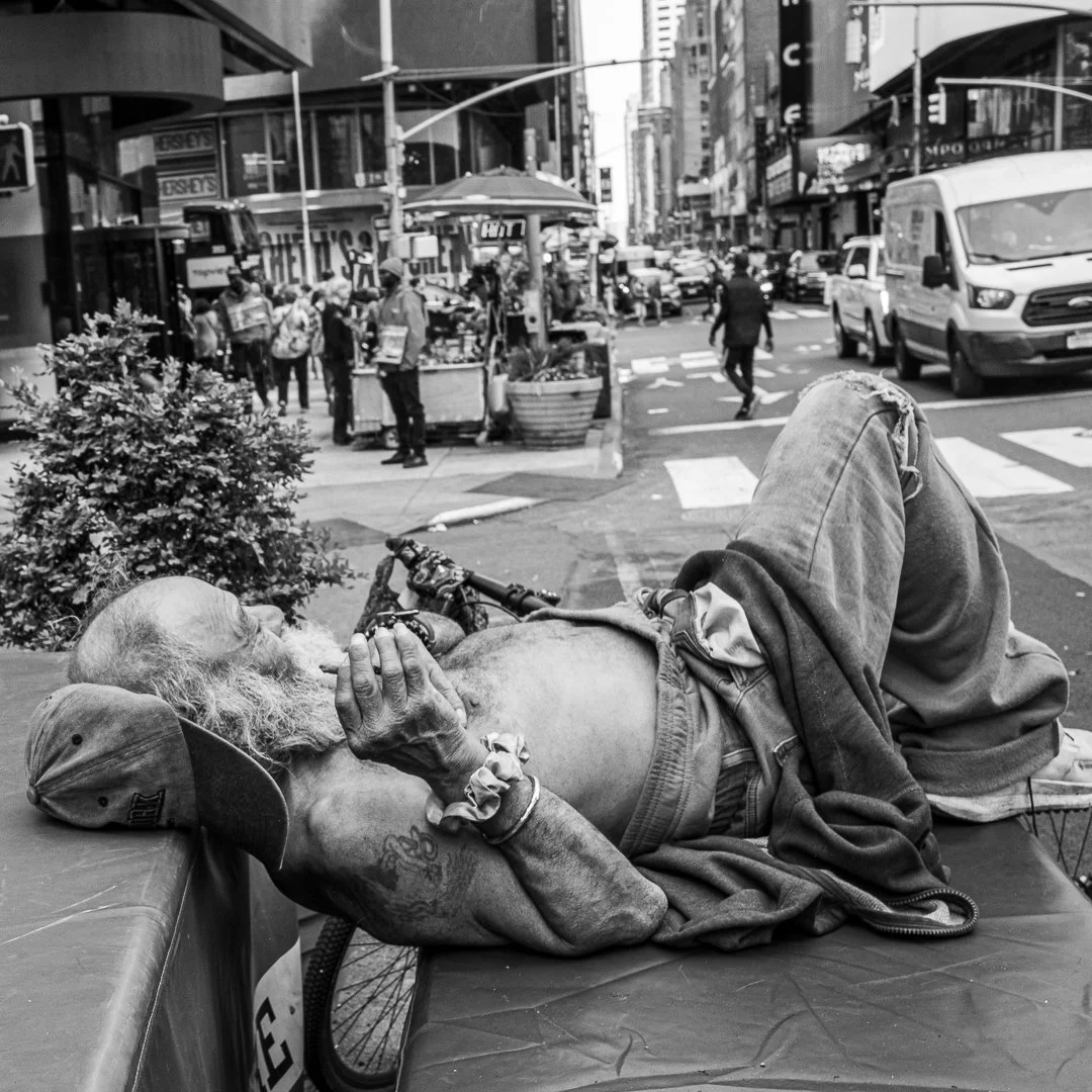 A bearded man rests comfortably on a bench beside his bicycle in a bustling city scene. People walk past, cars move through the street, and storefronts line the background, creating a contrast between his stillness and the surrounding motion.
