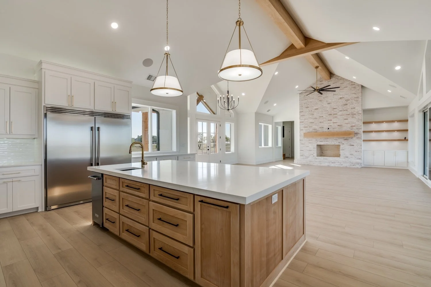 Open-concept kitchen and living room with a large island, white cupboards, stainless steel refrigerator, wooden beams, brick fireplace, built-in shelves, and multiple windows providing natural light, built by Lowrance Signature Homes.