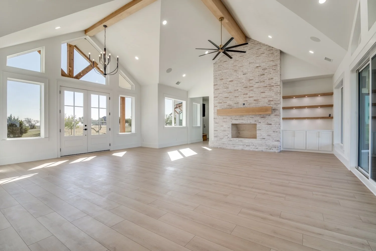 Empty living room with large windows, wooden floors, a brick fireplace, a ceiling fan, built-in shelves, and a chandelier